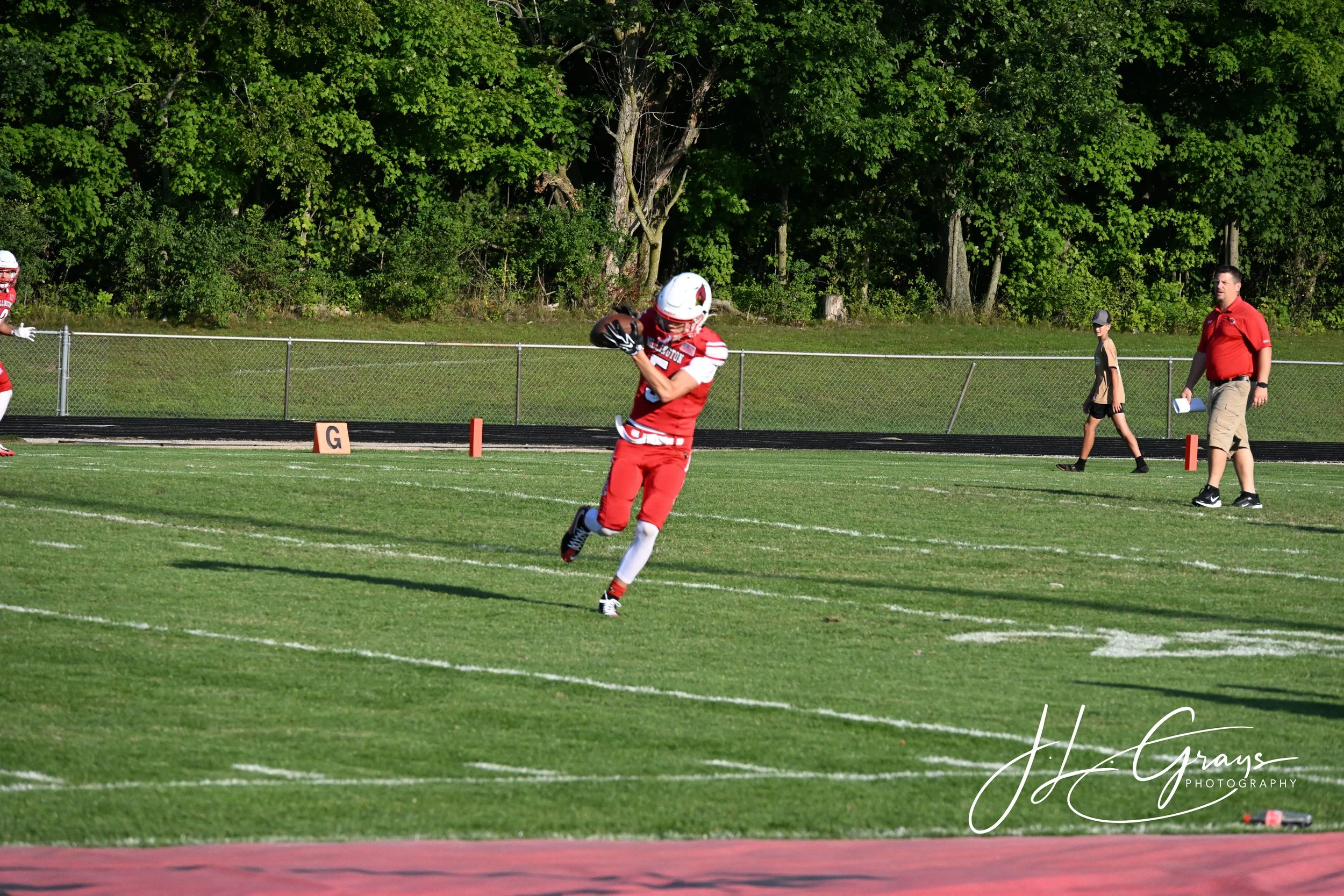 A football player in a red uniform and helmet catches a football on a green field. There are coaches and officials in the background near a chain-link fence and trees.