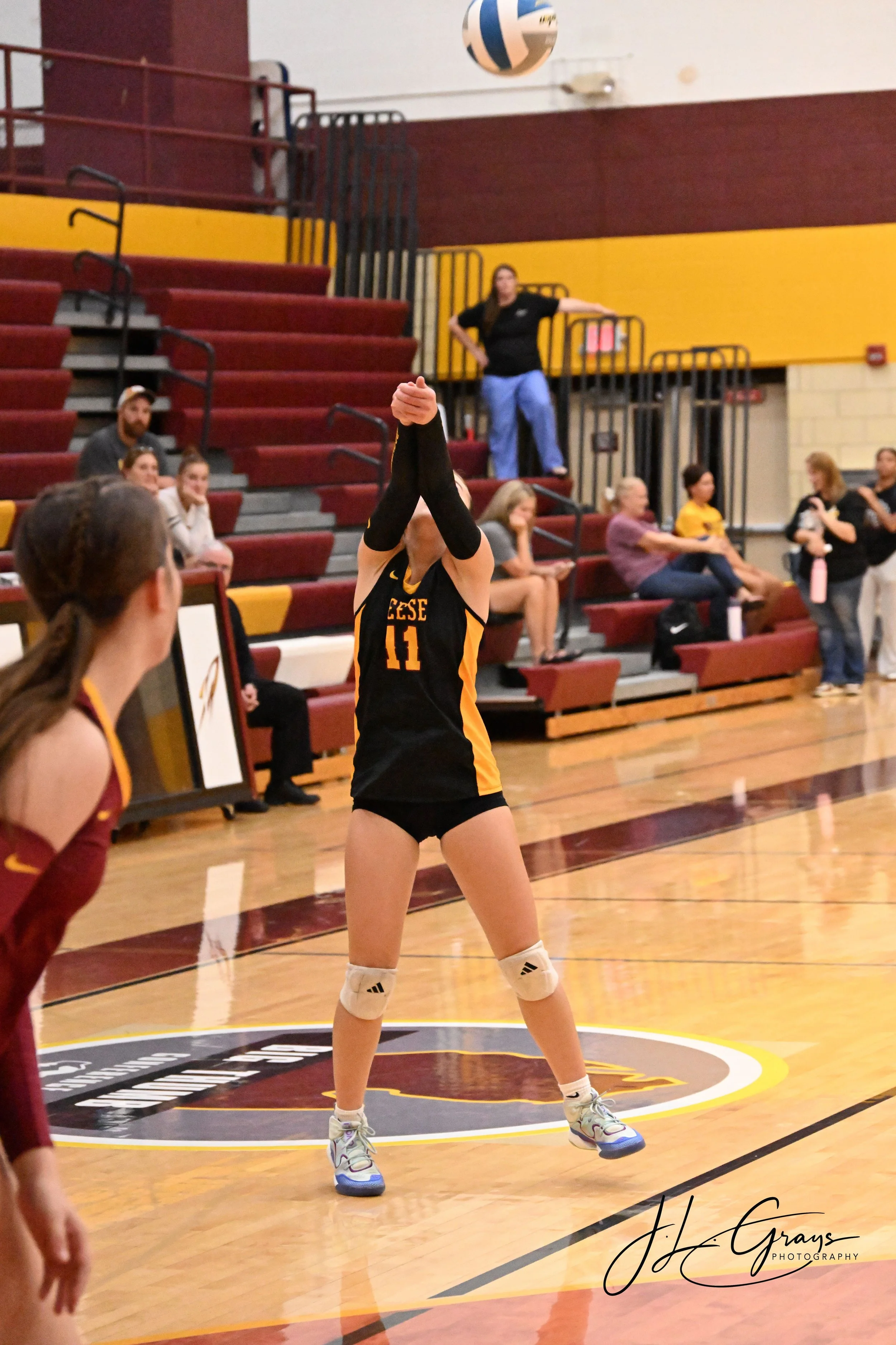 A volleyball player in black and yellow uniform preparing to serve during a game in a gym with spectators seated on red and yellow bleachers.