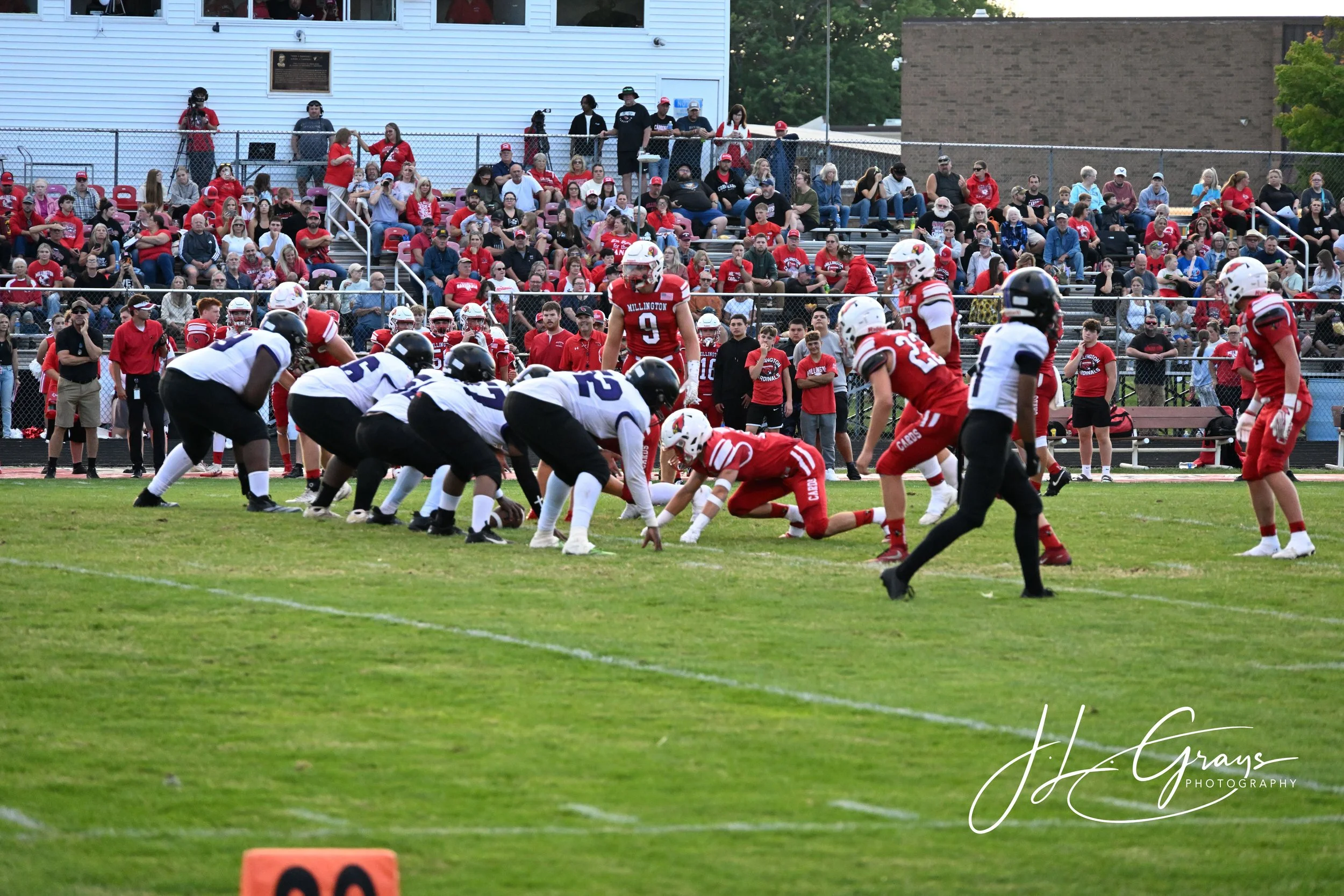 A high school football game in progress with players on the field and spectators in the stands, many wearing red and black. The players are lined up, ready for the next play, and the crowd watches attentively.