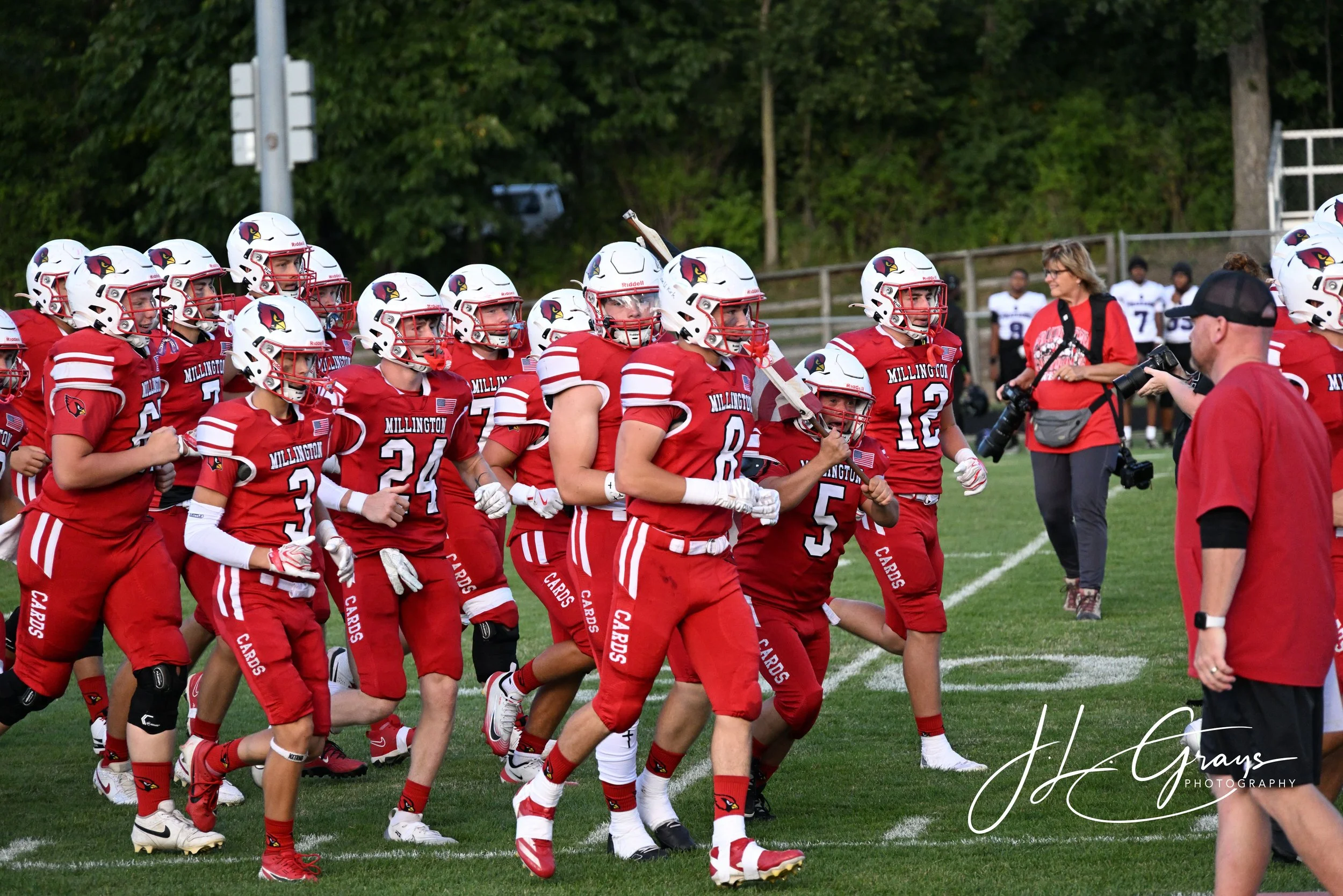 High school football team in red uniforms and white helmets with a cardinal logo, running onto the field for a game, with a coach and other players in the background.