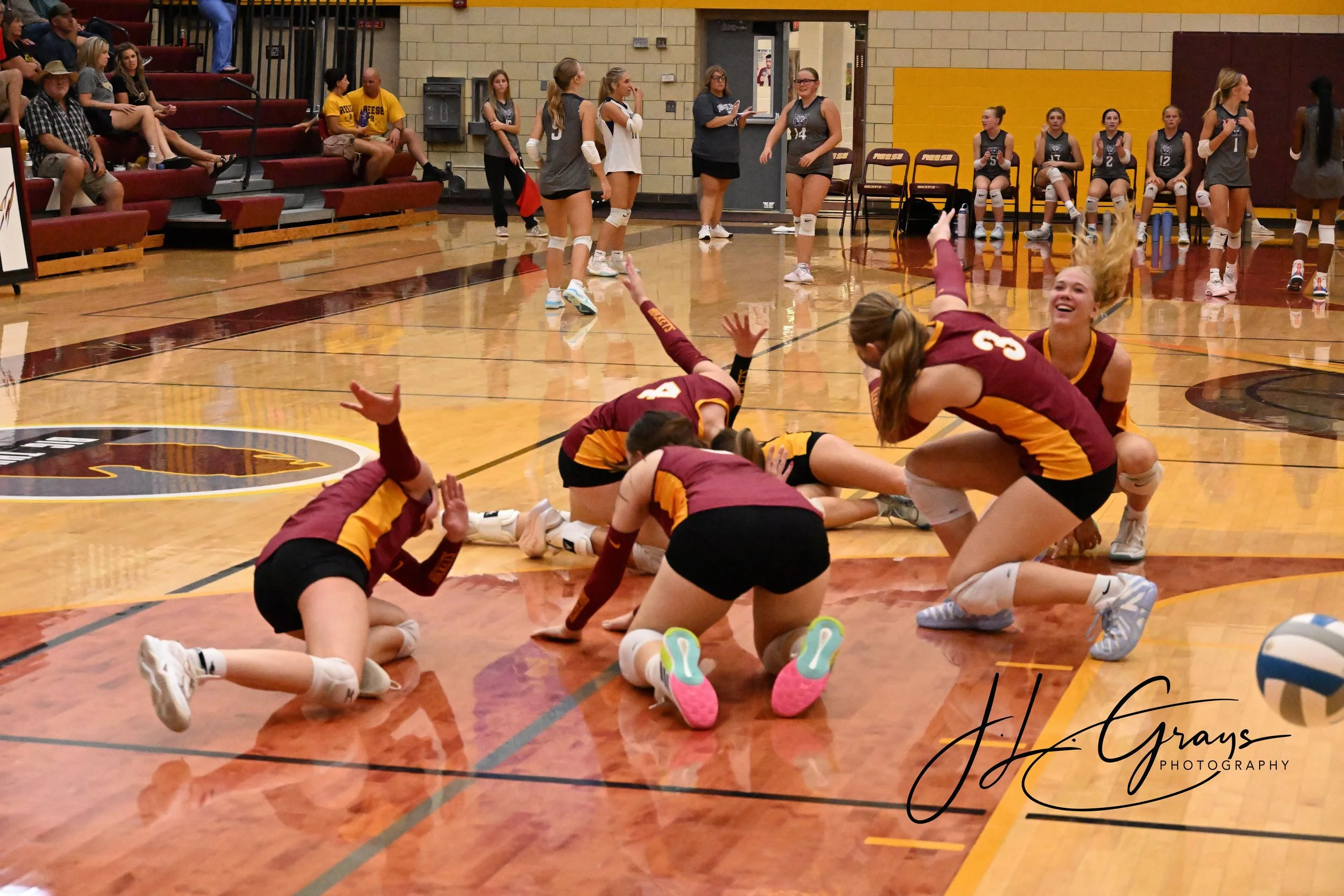 A volleyball game in progress with a female team in maroon and gold uniforms on the court, some players on the floor and others standing, celebrating or preparing for play. Spectators are seated on the bleachers in the background.