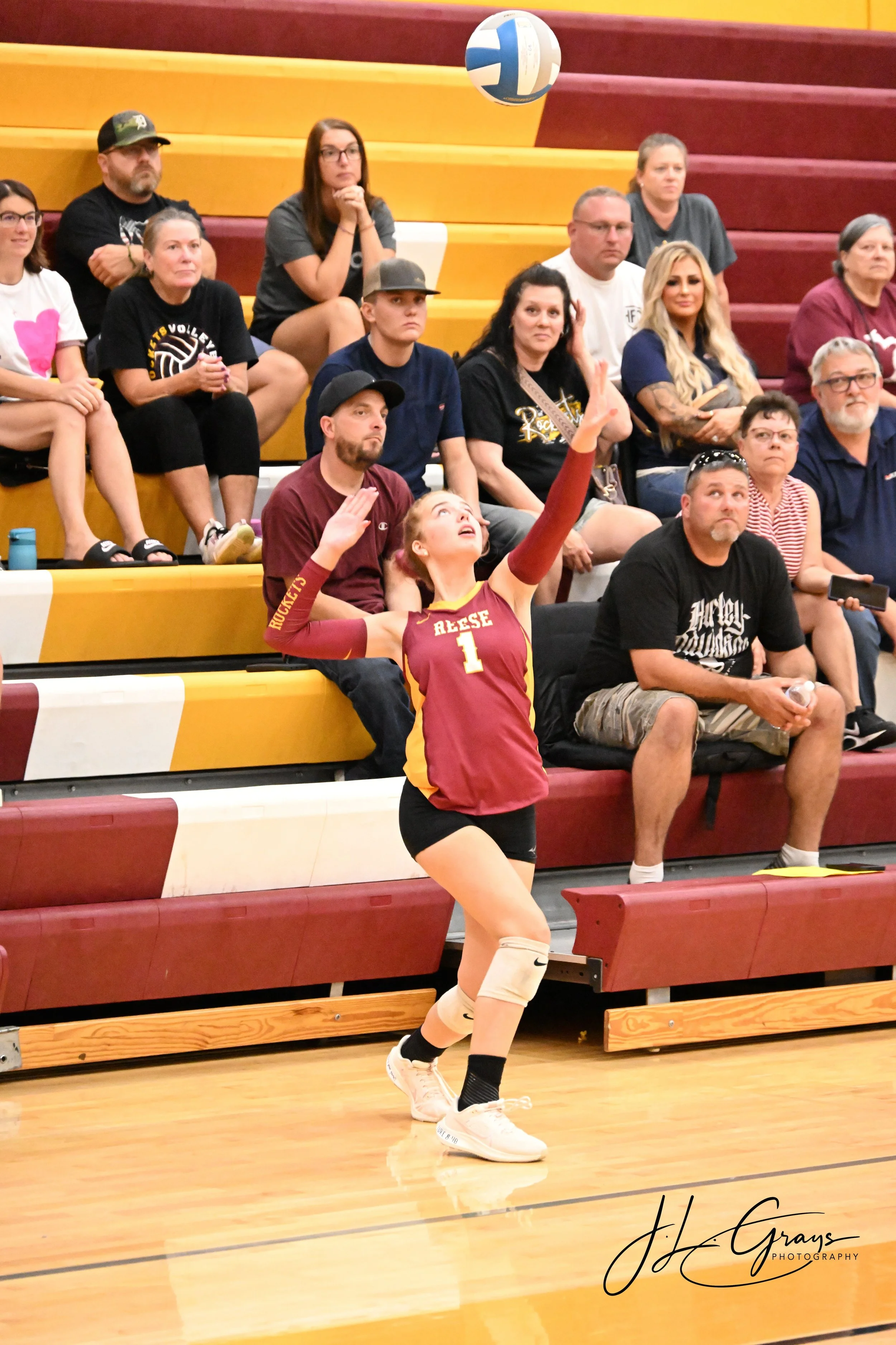 A female volleyball player in a maroon and yellow uniform is reaching up to hit a volleyball during a game. She is wearing knee-high socks and athletic shoes, with her hair tied back. Spectators sit on yellow and red bleachers in the background, watc