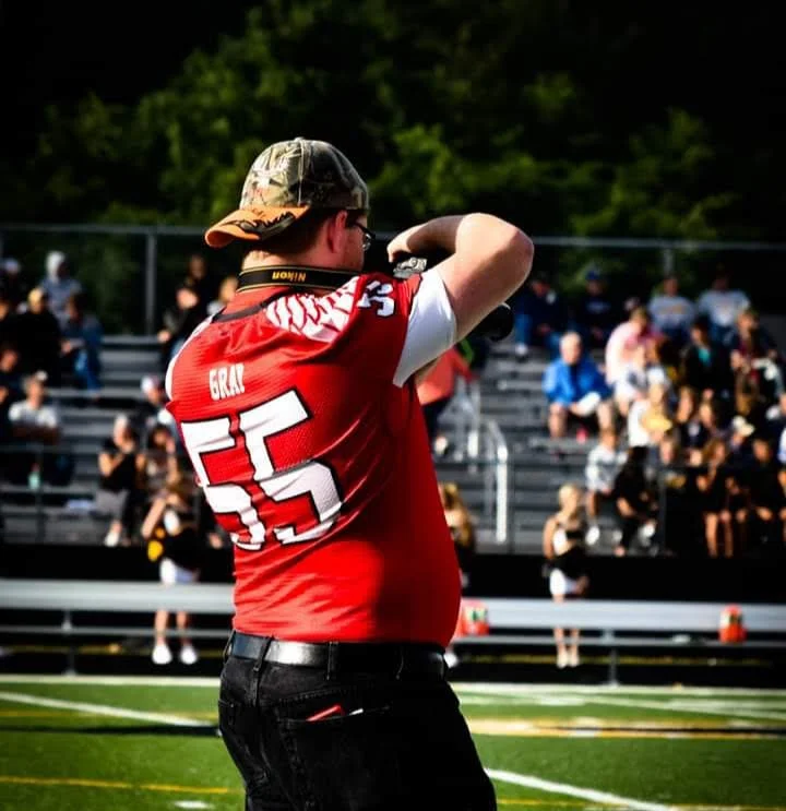 A person wearing a red sports jersey with the number 55, a camouflage cap, and glasses, on a football field taking a photo or video with their phone, with spectators in the background.