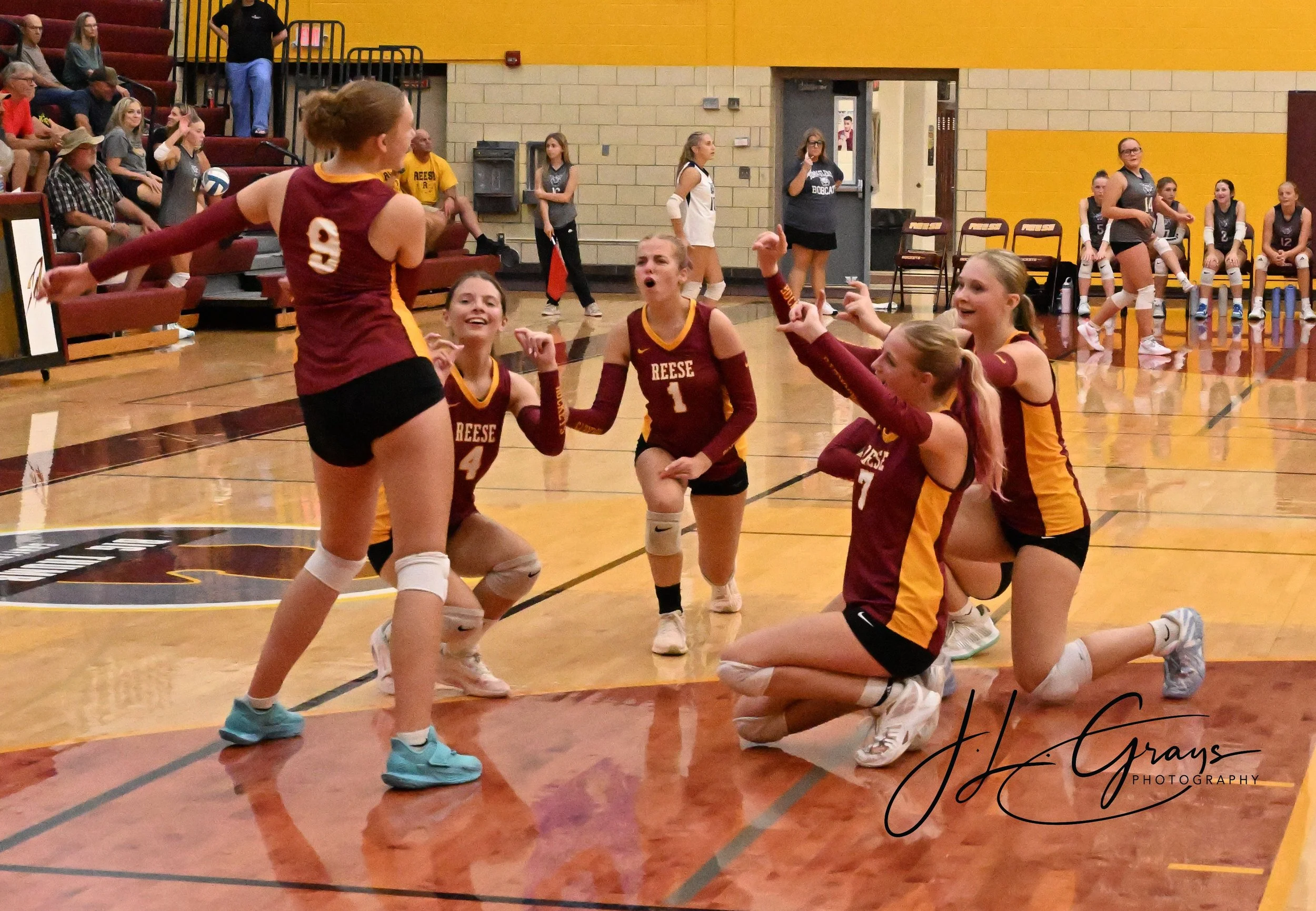 A volleyball game features players from Reese, with some kneeling on the court and others standing, celebrating or talking. Spectators and team benches are visible in the background inside a gymnasium with yellow and beige walls.