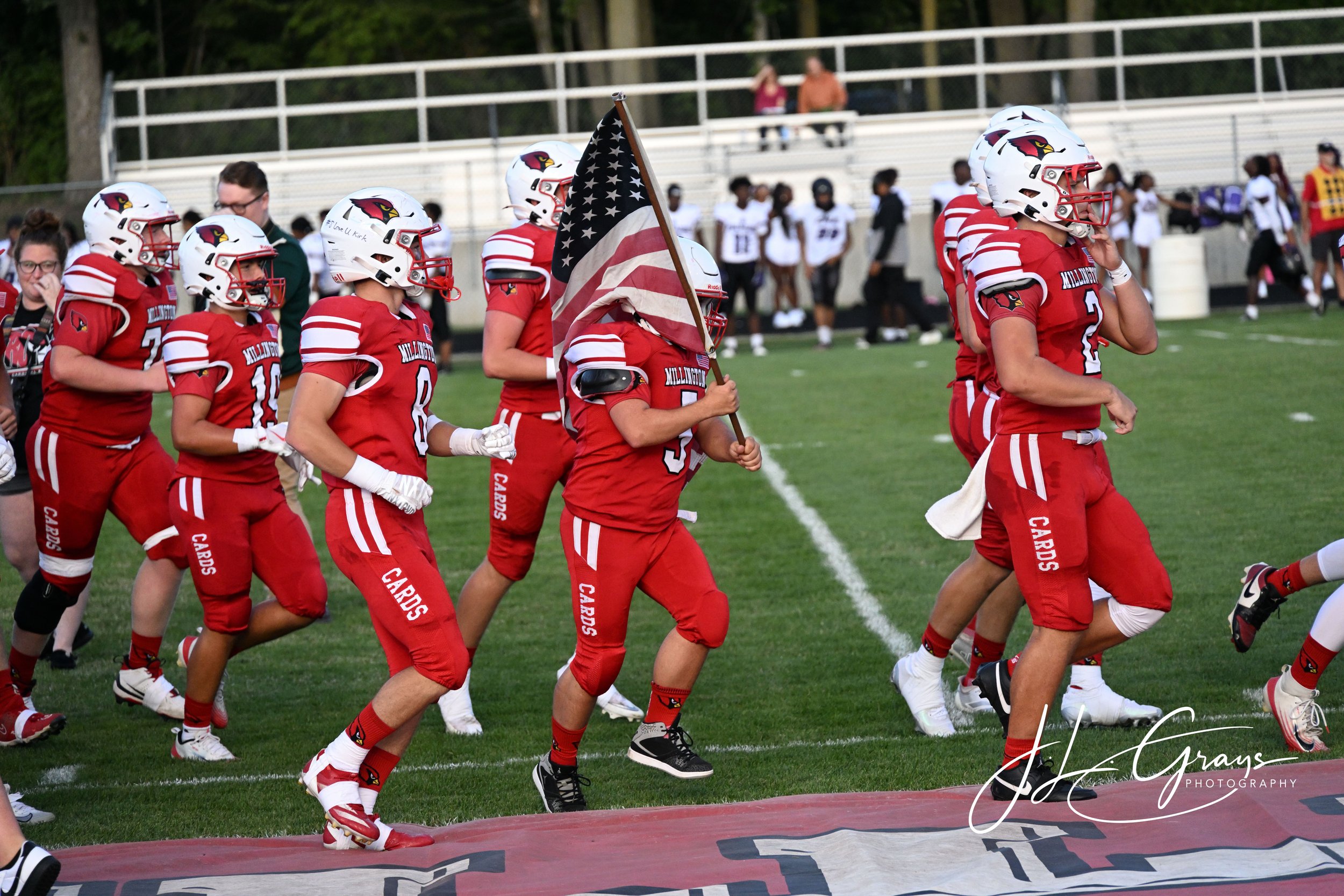 A group of football players in red uniforms and white helmets, some holding an American flag, running onto the field during a game.