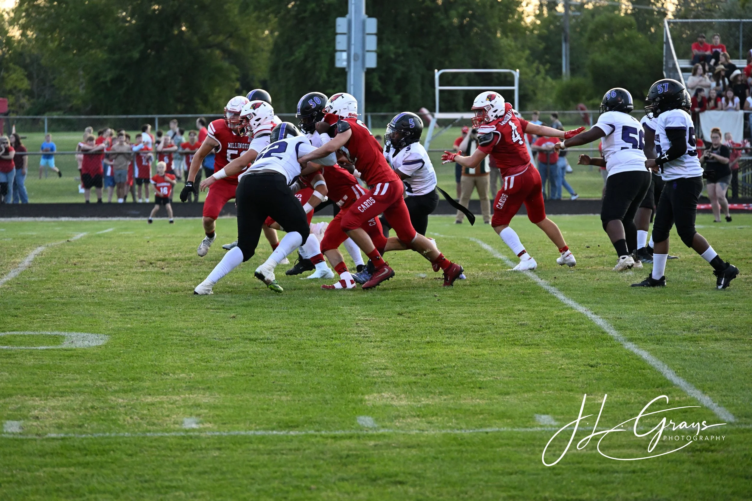 A youth football game with players wearing red and white uniforms on a grassy field, some players engaged in blocking and tackling, with spectators watching from the sidelines and bleachers.