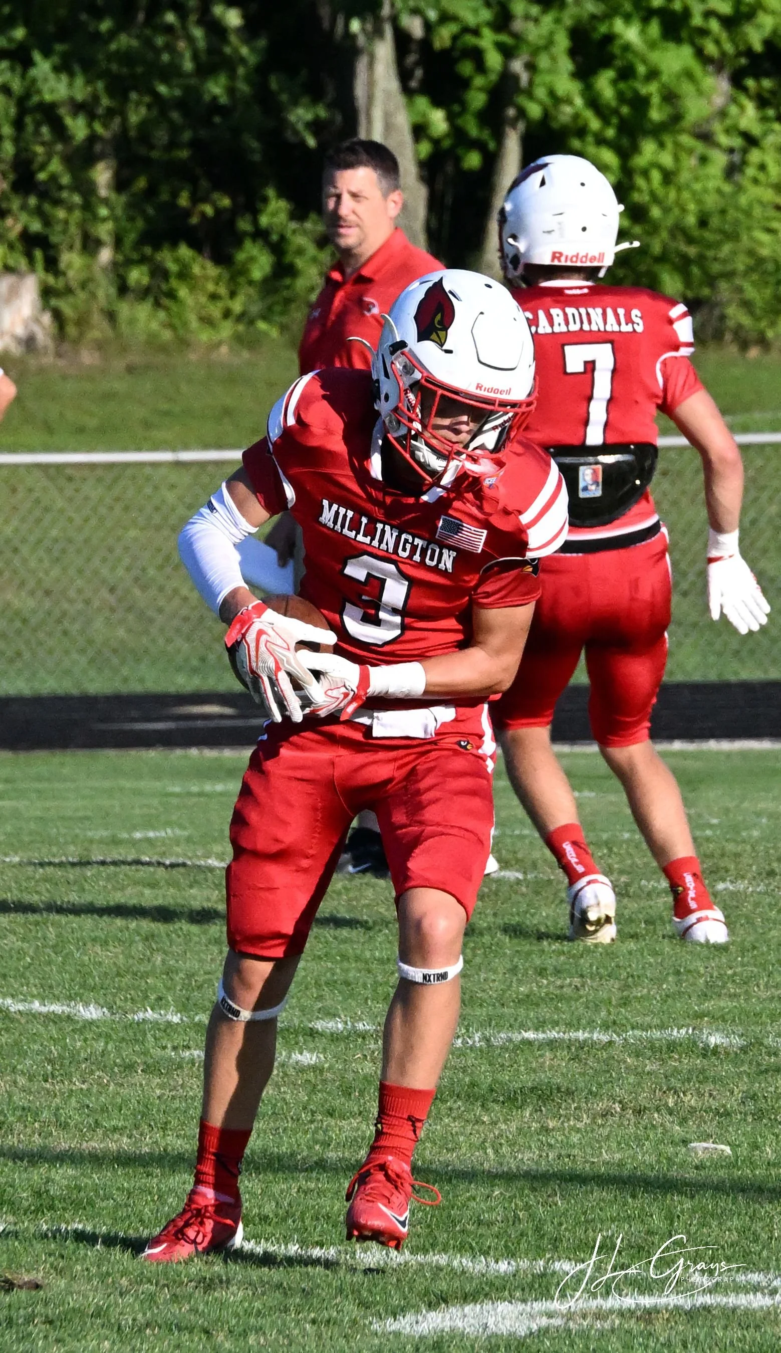 American football player in red uniform with number 3 and team name Millington running on football field, holding football, with two teammates behind him wearing red jerseys, white helmets, and red pants, and a coach in the background watching.