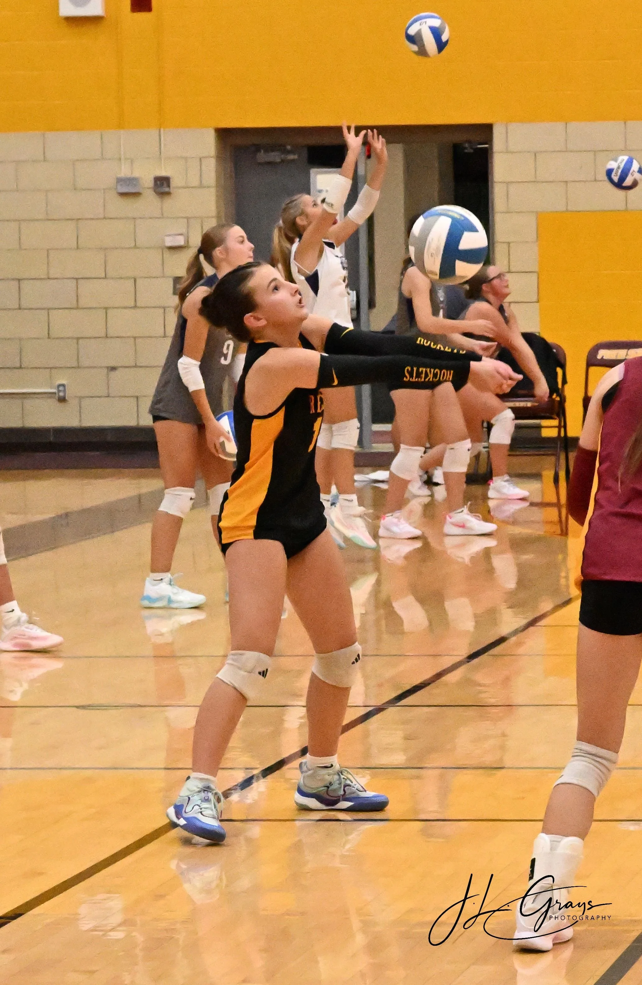 Girls volleyball team practicing in gym, one girl in black and yellow jersey preparing to pass, other team members standing behind her, volleyballs in mid-air.