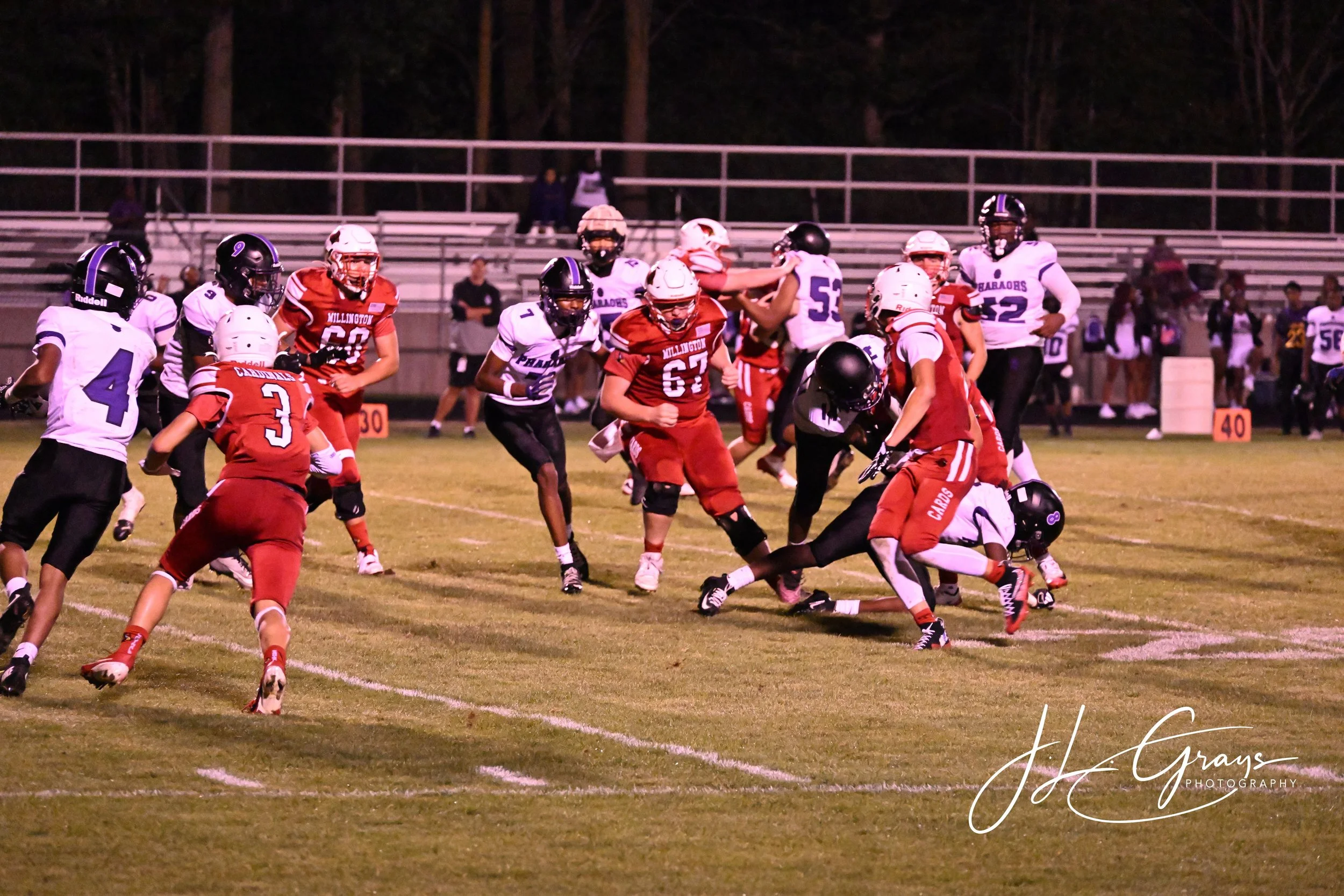 Youth football game at night with players in white and red uniforms on a grassy field with bleachers and trees in the background.