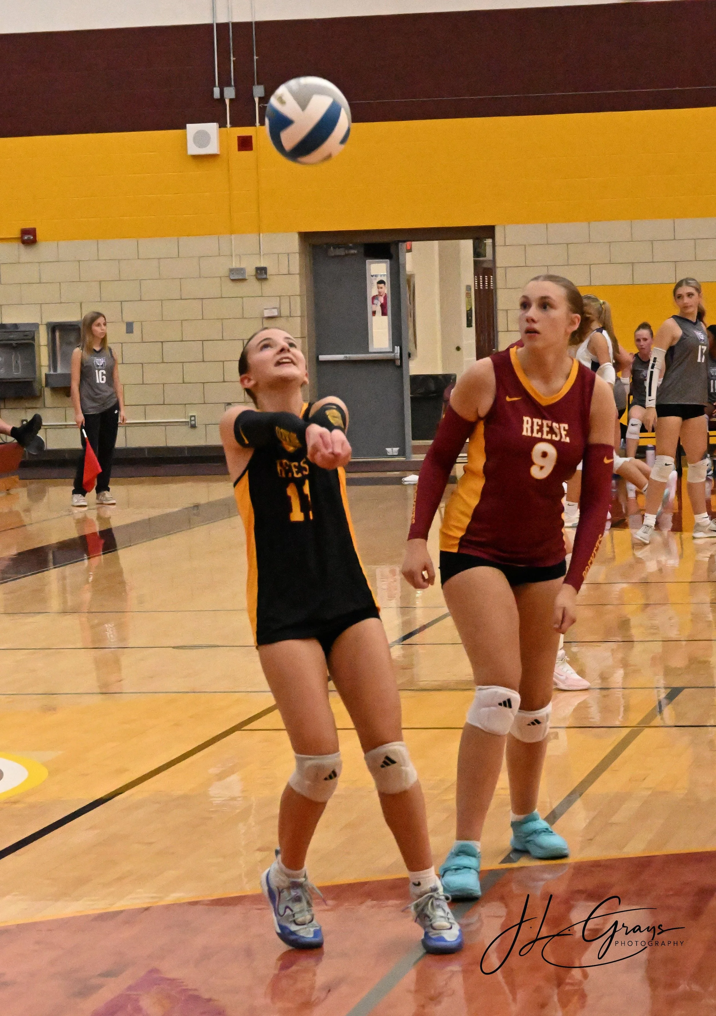 Two female volleyball players in action during a game in an indoor gymnasium, with other players and a referee in the background.
