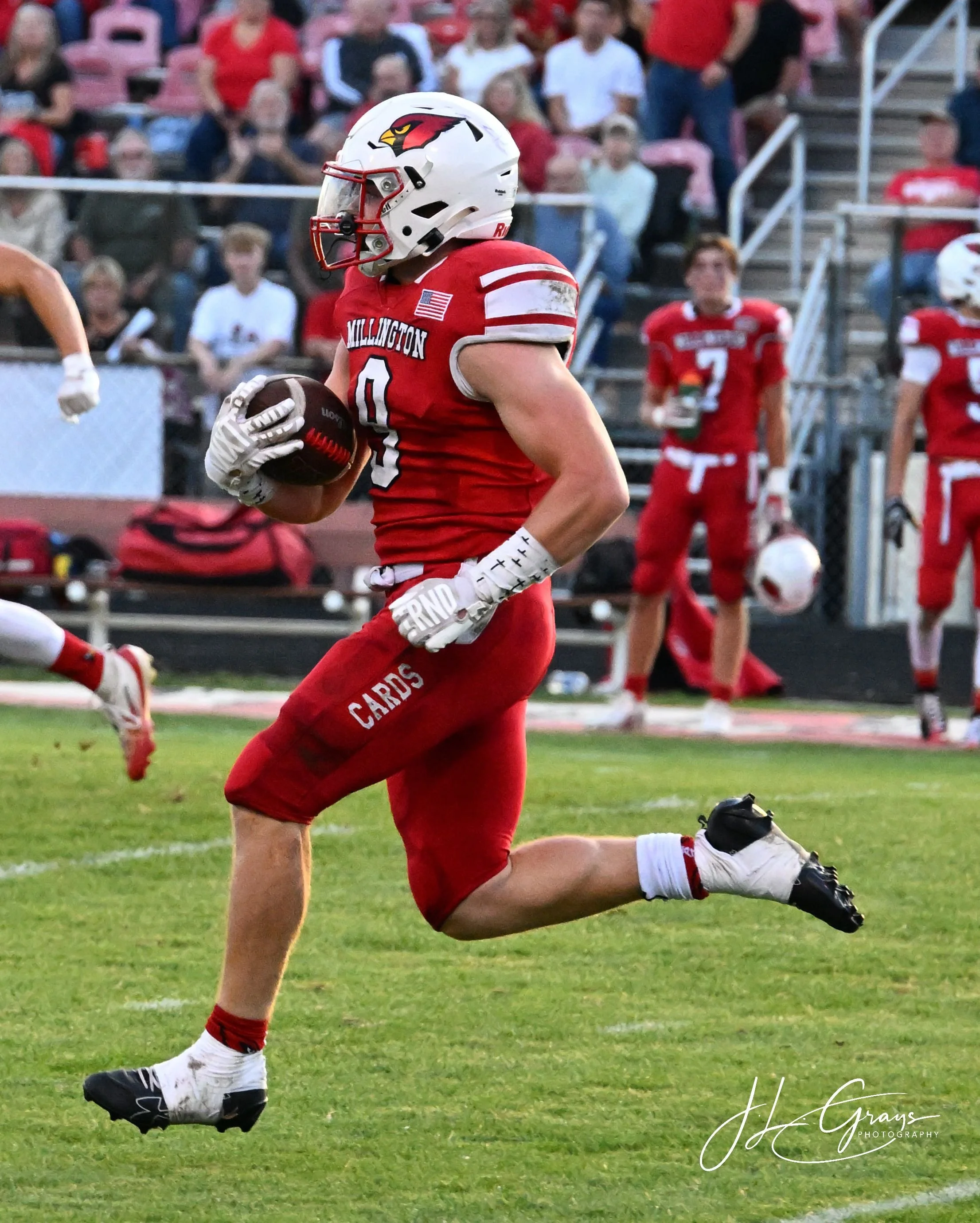 American football player in a red uniform with white helmet, running on the field holding a football, crowd in the stands in the background.