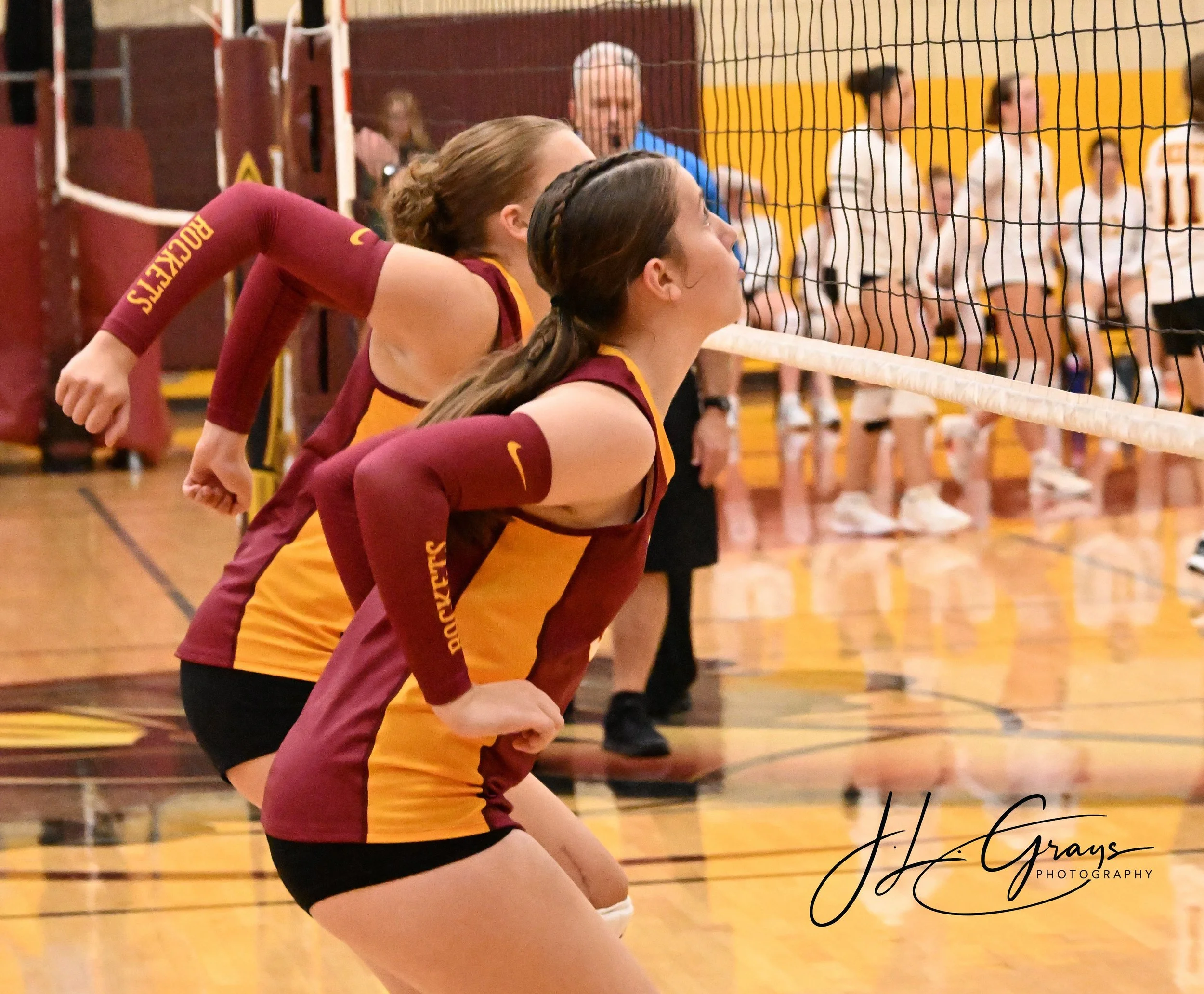 A group of women in volleyball uniforms kneeling on a gym floor, preparing for serve or receive, with volleyball net in the foreground and spectators in the background.