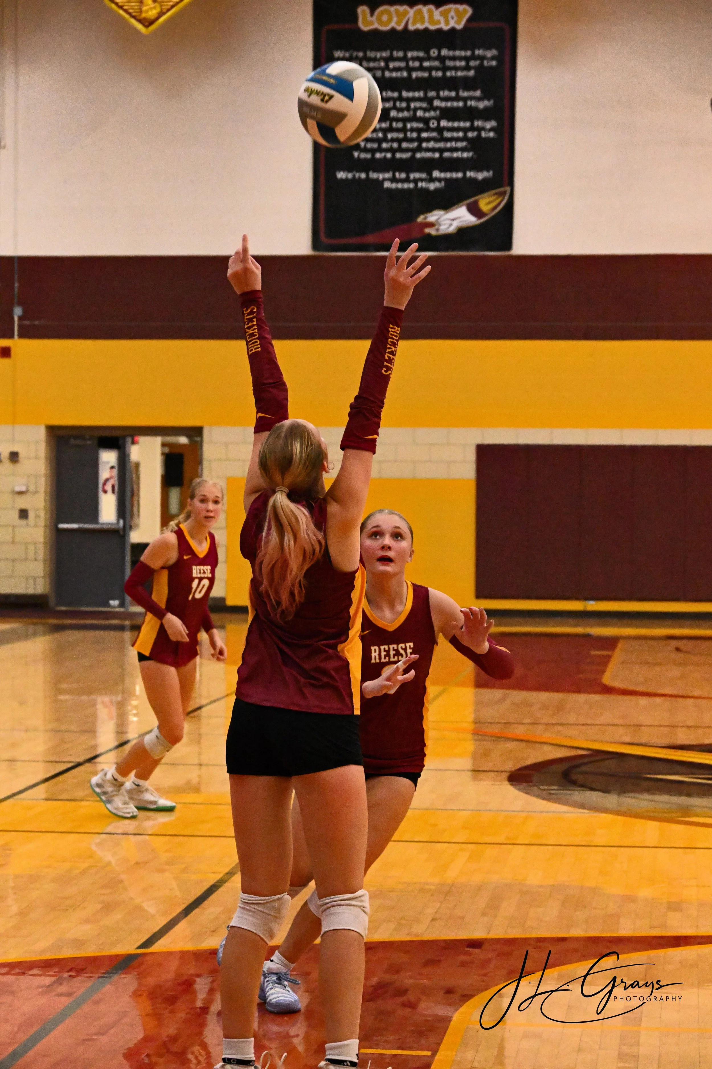 A volleyball game is taking place in a gym. A player in a maroon and yellow uniform with 'RESEE' on the front is preparing to pass the ball in the air. Two other players in similar uniforms, one with number 10, are visible in the background. The gym 