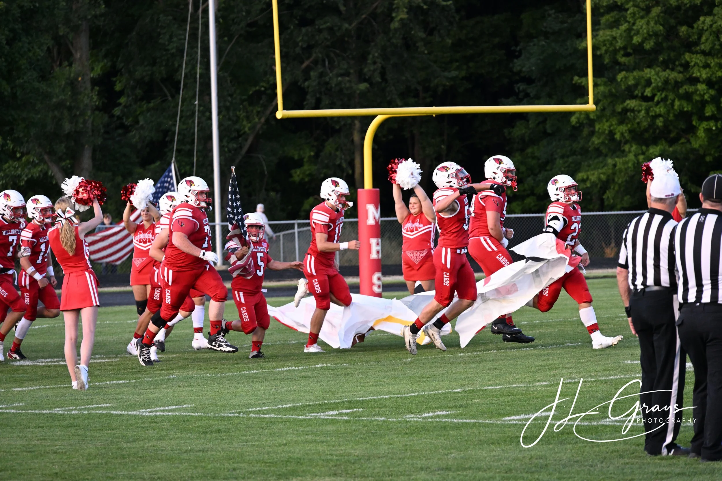 A group of high school football players in red uniforms and white helmets running onto the field with cheerleaders holding pom-poms and American flags. The scene is set at a football stadium with a goalpost and trees in the background.