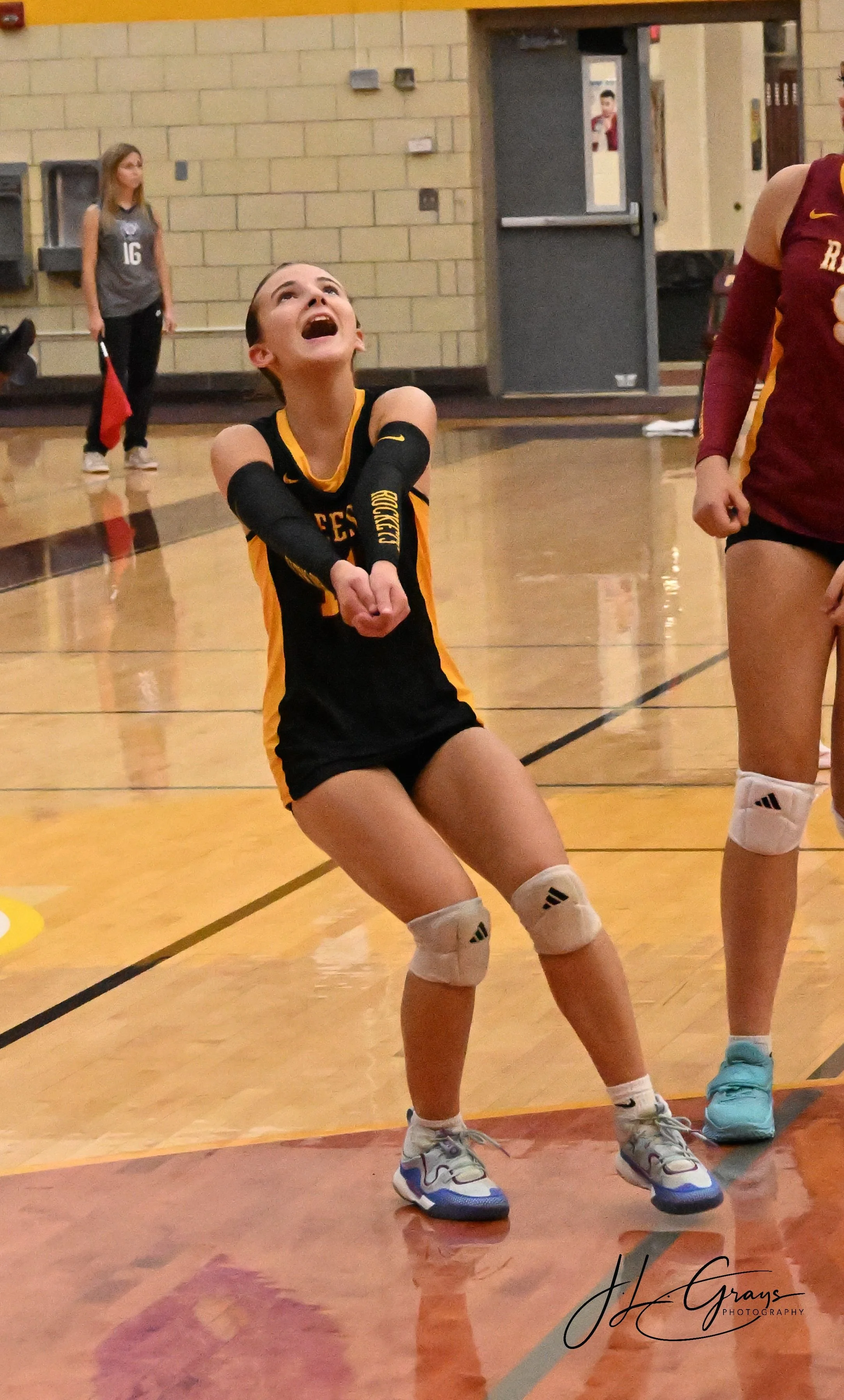 A female volleyball player in a black and yellow uniform appears to be falling to her knees with her mouth open, looking upward, on an indoor volleyball court. She is wearing knee pads and sneakers. In the background, another player in a maroon unifo