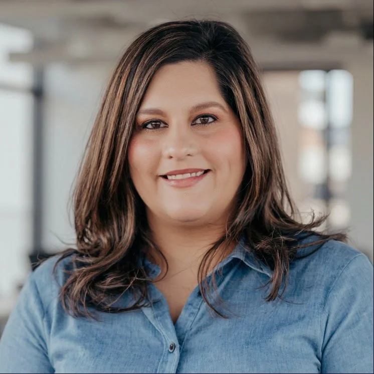 Portrait of a woman with brown hair in a denim shirt, smiling, indoors with blurred background.