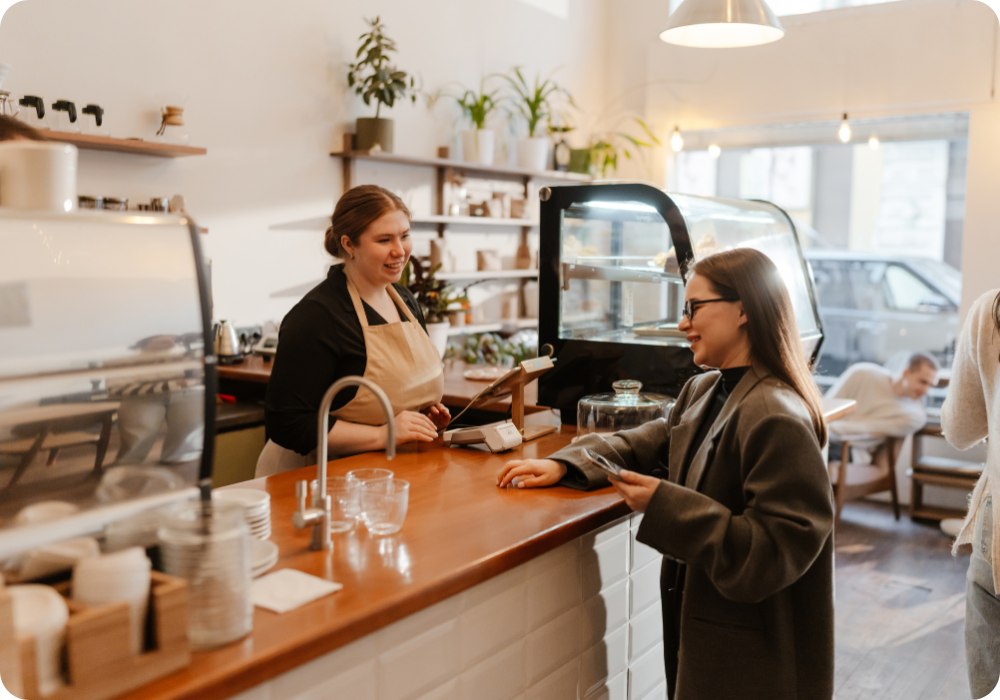 Young woman interacts with barista at cafe counter, smiling, as a customer pays with a smartphone in a bright, modern coffee shop.