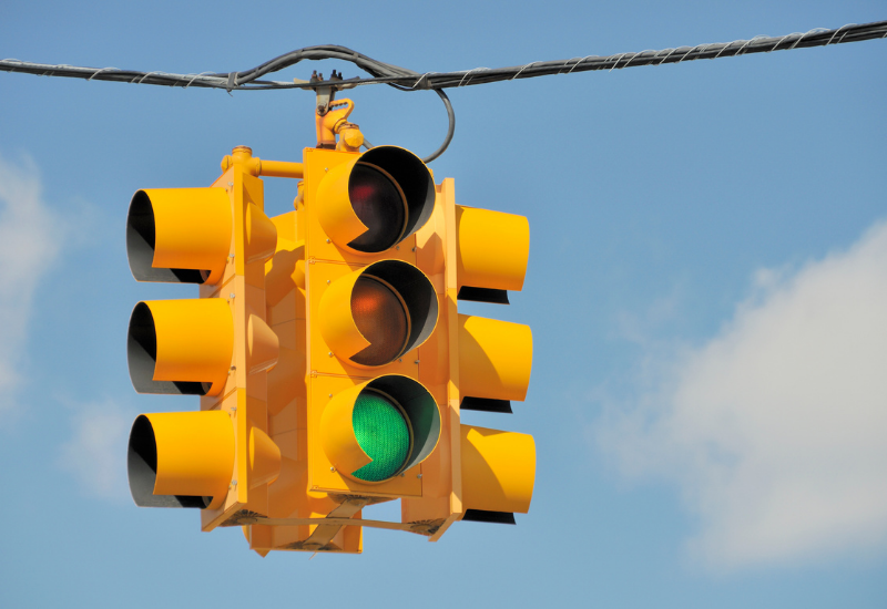A yellow traffic light with the green light illuminated, hanging above a road with a blue sky and clouds in the background.