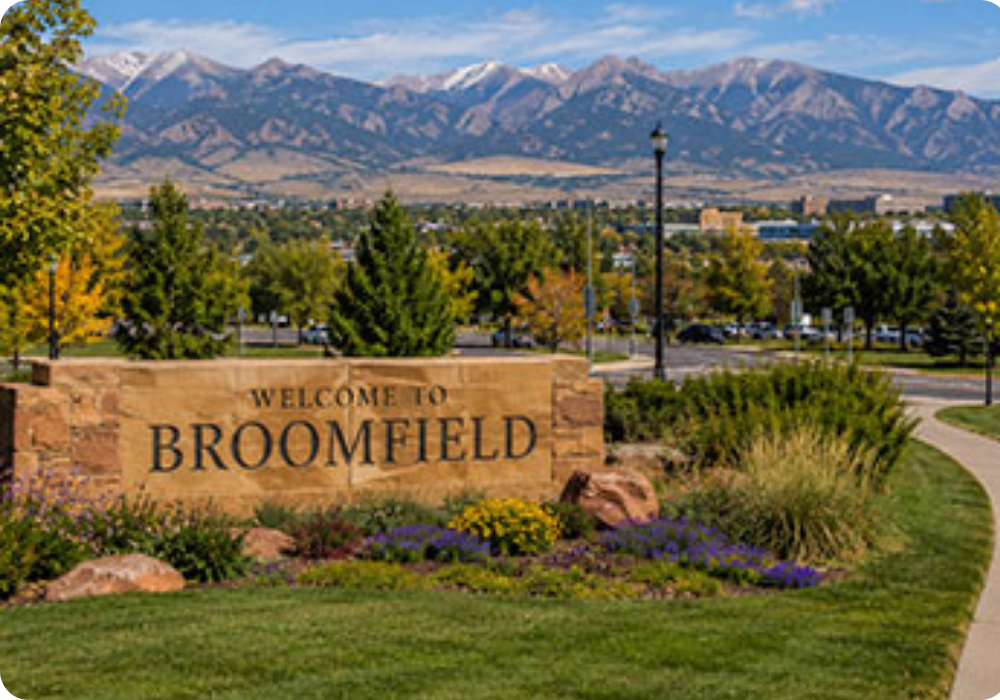 A sign reading 'Welcome to Broomfield' situated in a landscaped area with flowers and bushes, with a backdrop of trees, streetlights, and snow-capped mountains under a partly cloudy sky.