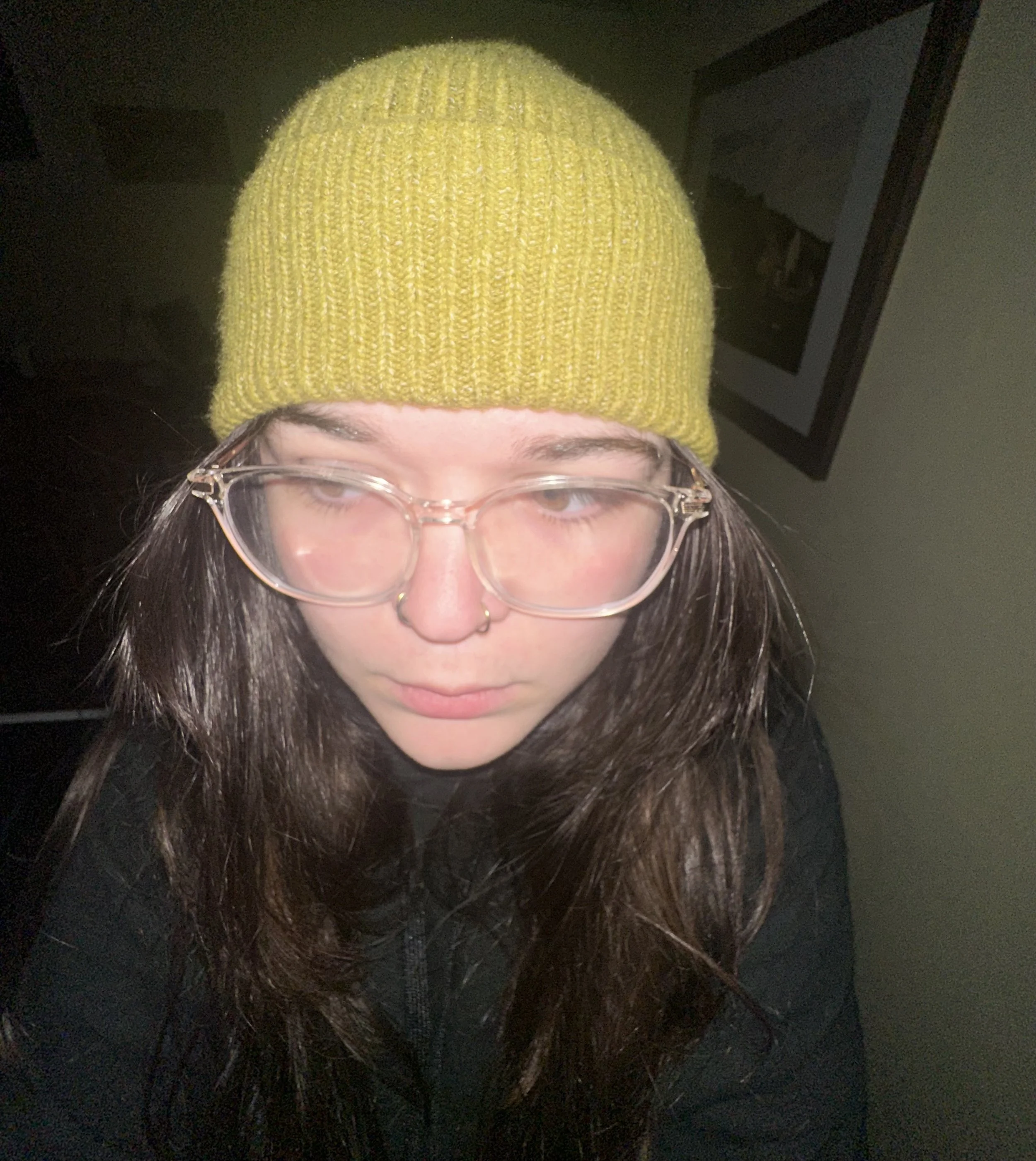 Person wearing a yellow knit beanie and clear glasses, with long brown hair, looking downward. There is a framed picture on the wall in the background.