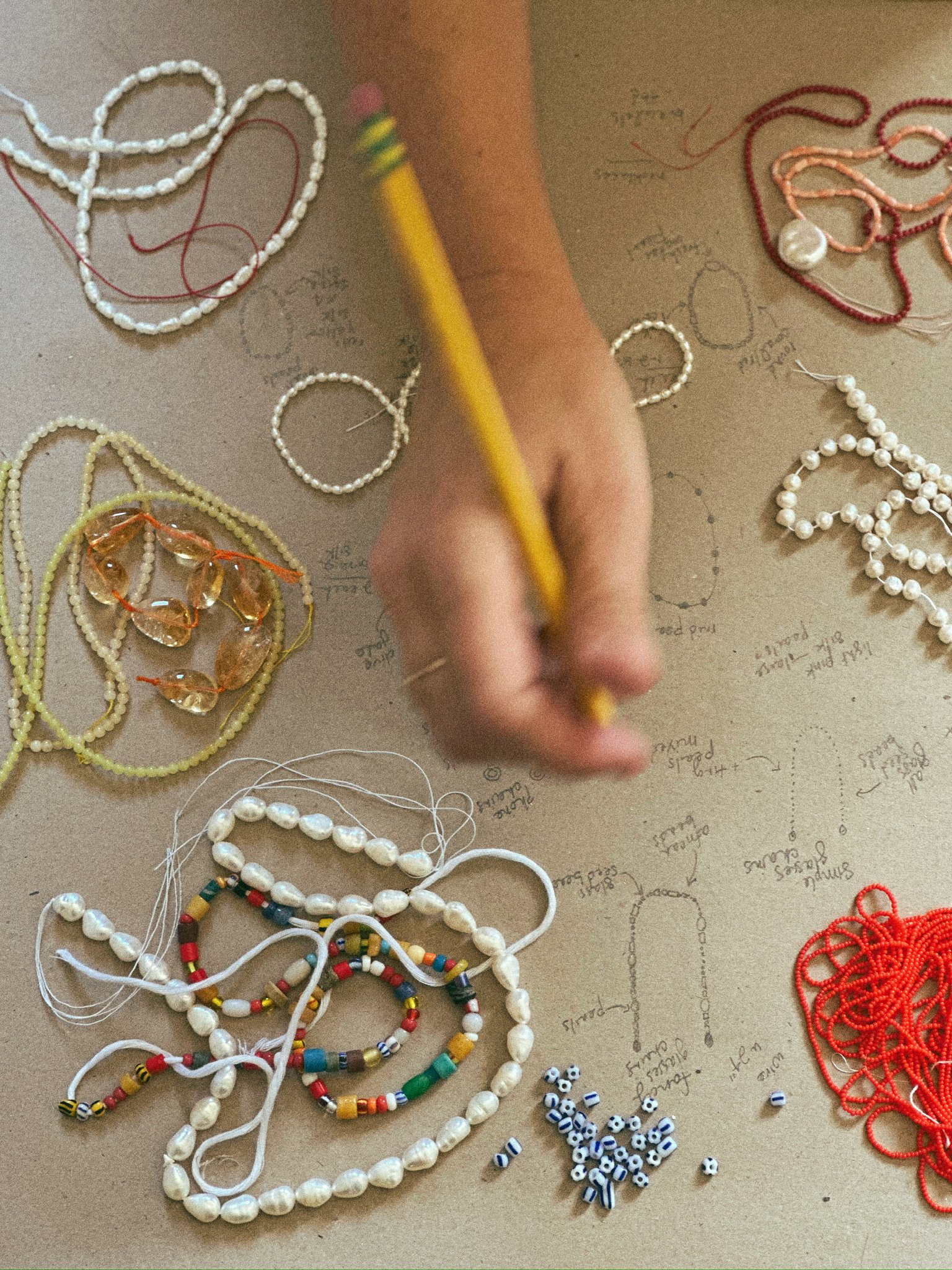 A person's hand with a yellow pencil working on a craft project involving various handmade necklaces and beads, with handwritten notes and sketches on brown paper in the background.