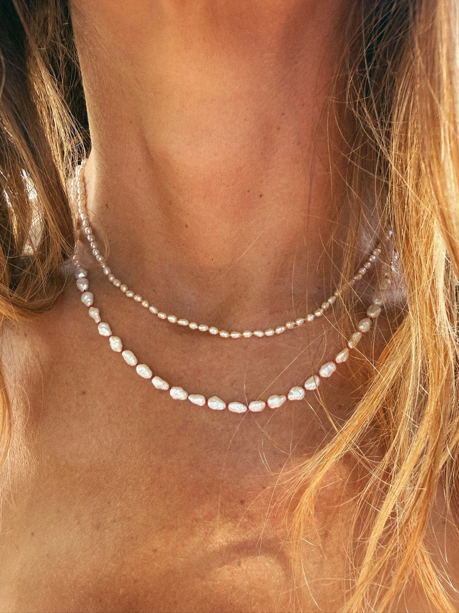 Small knotted pearl necklace and larger knotted pearl necklace shown worn together on person's neck, close up with hair and chest