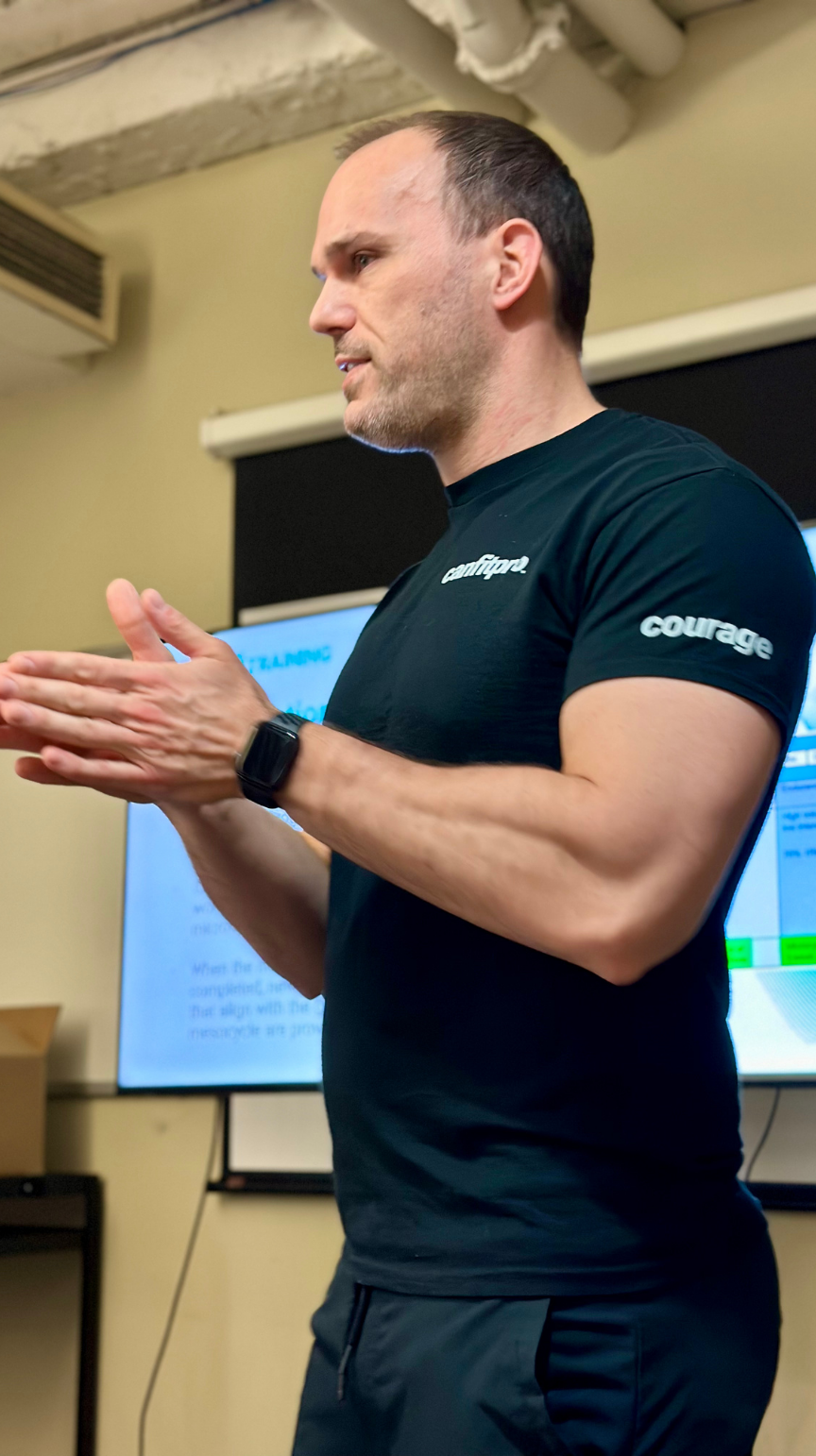 A man with short dark hair and a beard, wearing a black t-shirt with the words 'confidence' and 'courage' on it, standing in front of a presentation screen, gesturing with his hands during a presentation.