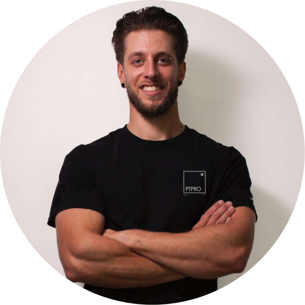 A smiling young man with a beard and short hair, wearing a black t-shirt with a logo, standing against a white wall with arms crossed.