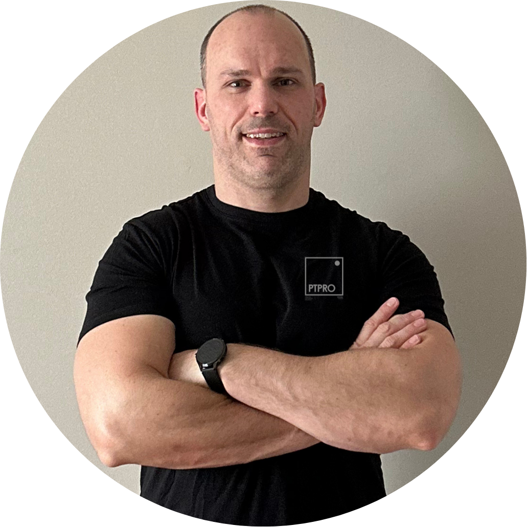 A man with a shaved head and light beard, dressed in a black t-shirt with a small logo, standing with arms crossed in front of a plain beige wall.