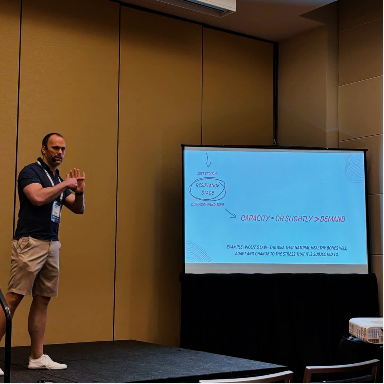 A man is giving a presentation in a conference room with a large screen displaying handwritten notes about resistance stage and capacity versus demand, with a few arrows and some text.