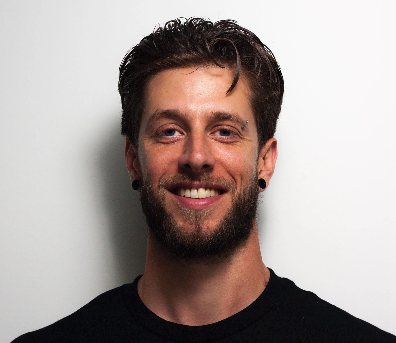 Close-up of a smiling man with dark hair, beard, and multiple piercings on his eyebrows and ears, standing against a plain gray background.