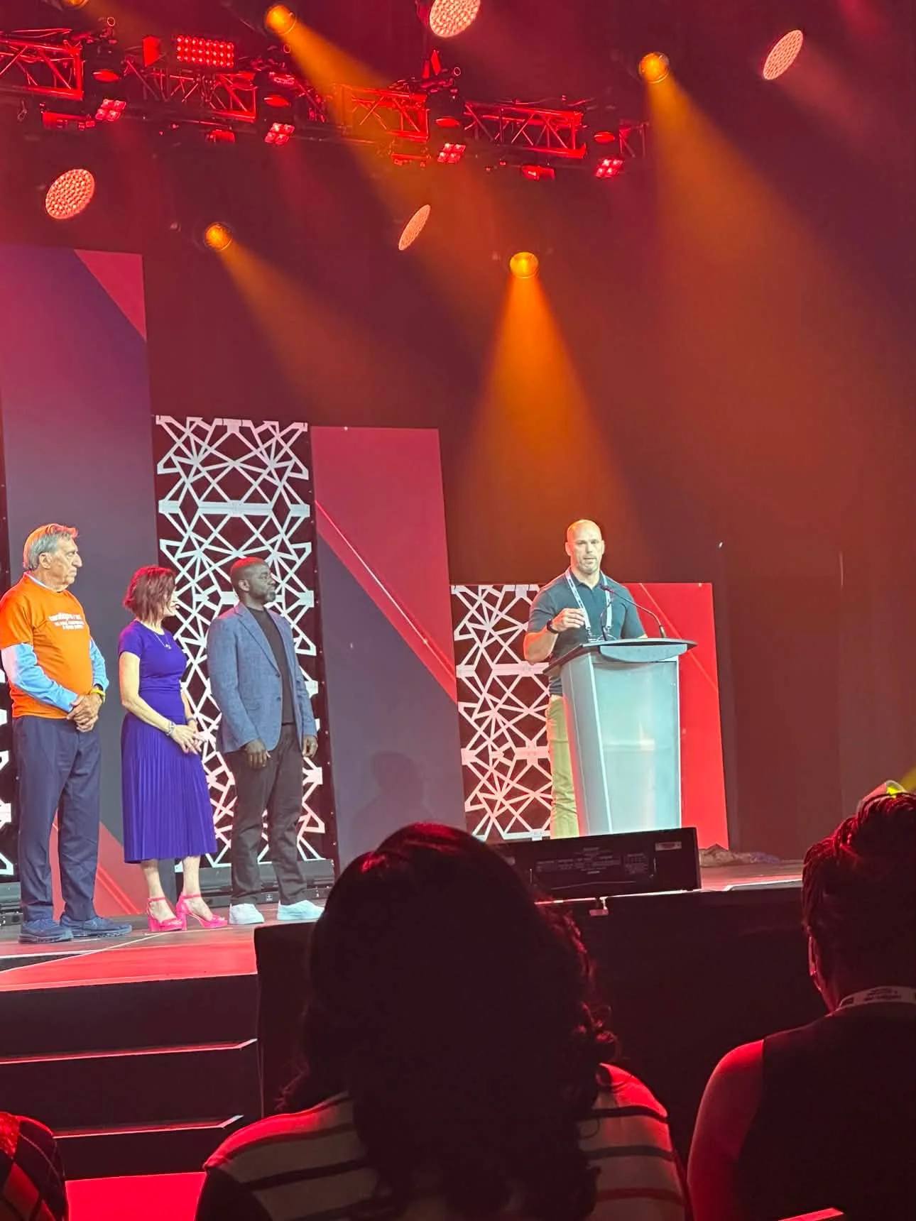 A group of five people on stage at a conference, with one person speaking at a podium and the others standing in a line behind him. The stage has red and black geometric backgrounds and bright lighting.