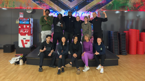 Group of ten students graduates posing for a photo in a fitness or dance studio with a colorful wall mural in the background. Seven people are standing in the back row and three are sitting in front. They are all smiling and looking at the camera.
