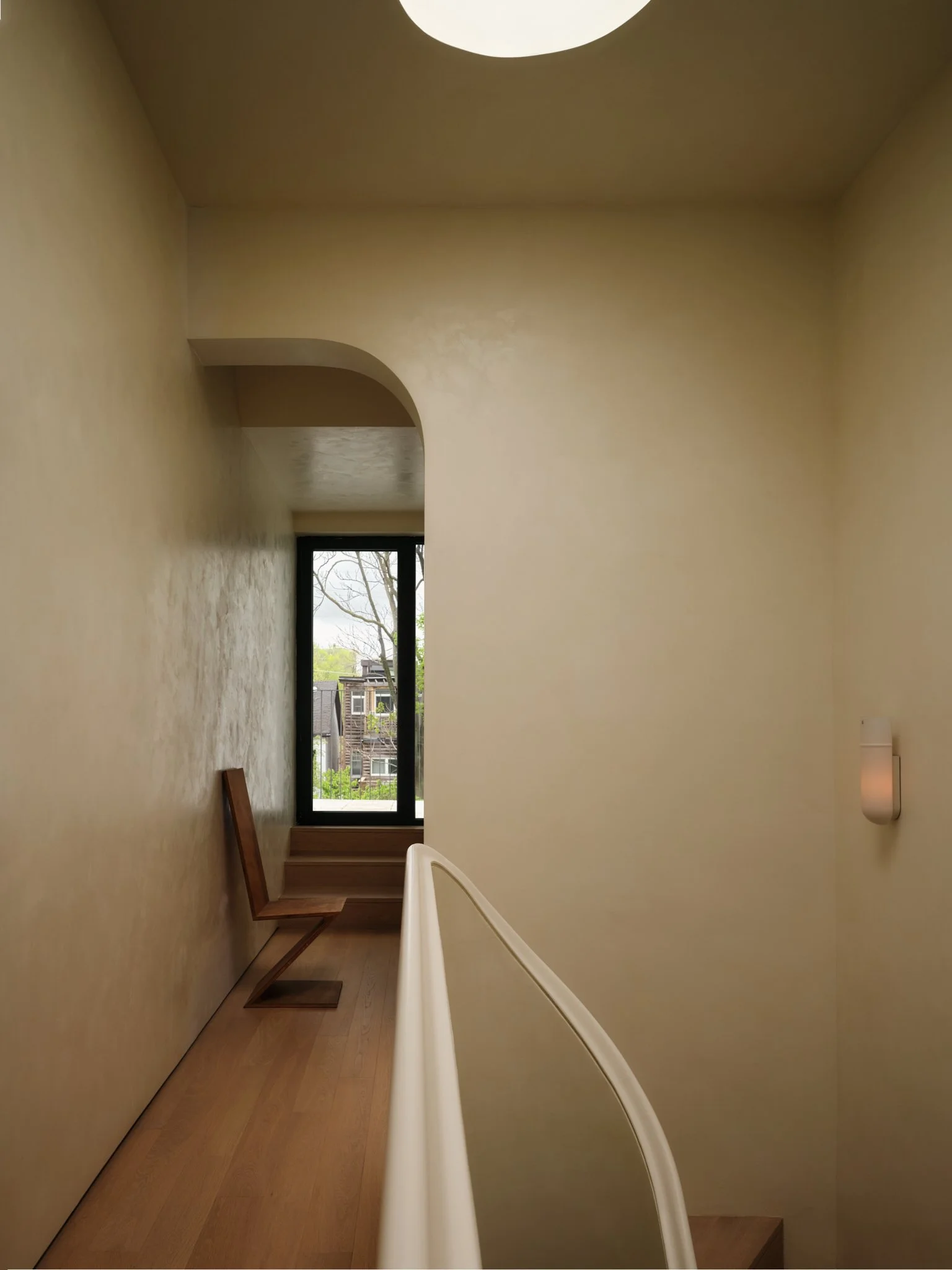 A hallway with wood flooring, beige walls, a window at the end showing trees and buildings outside, a wooden bench on the left, and a white curved handrail on the right side.
