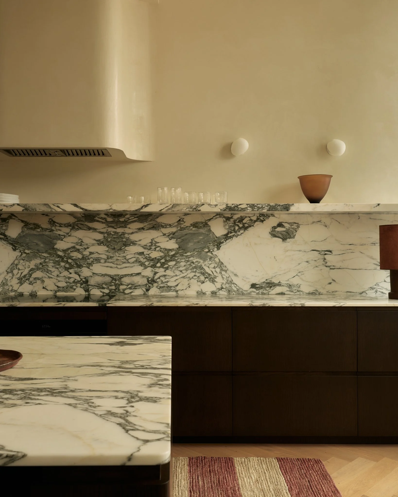 A kitchen with a marble countertop, wooden cabinets, and a red striped rug on the floor.