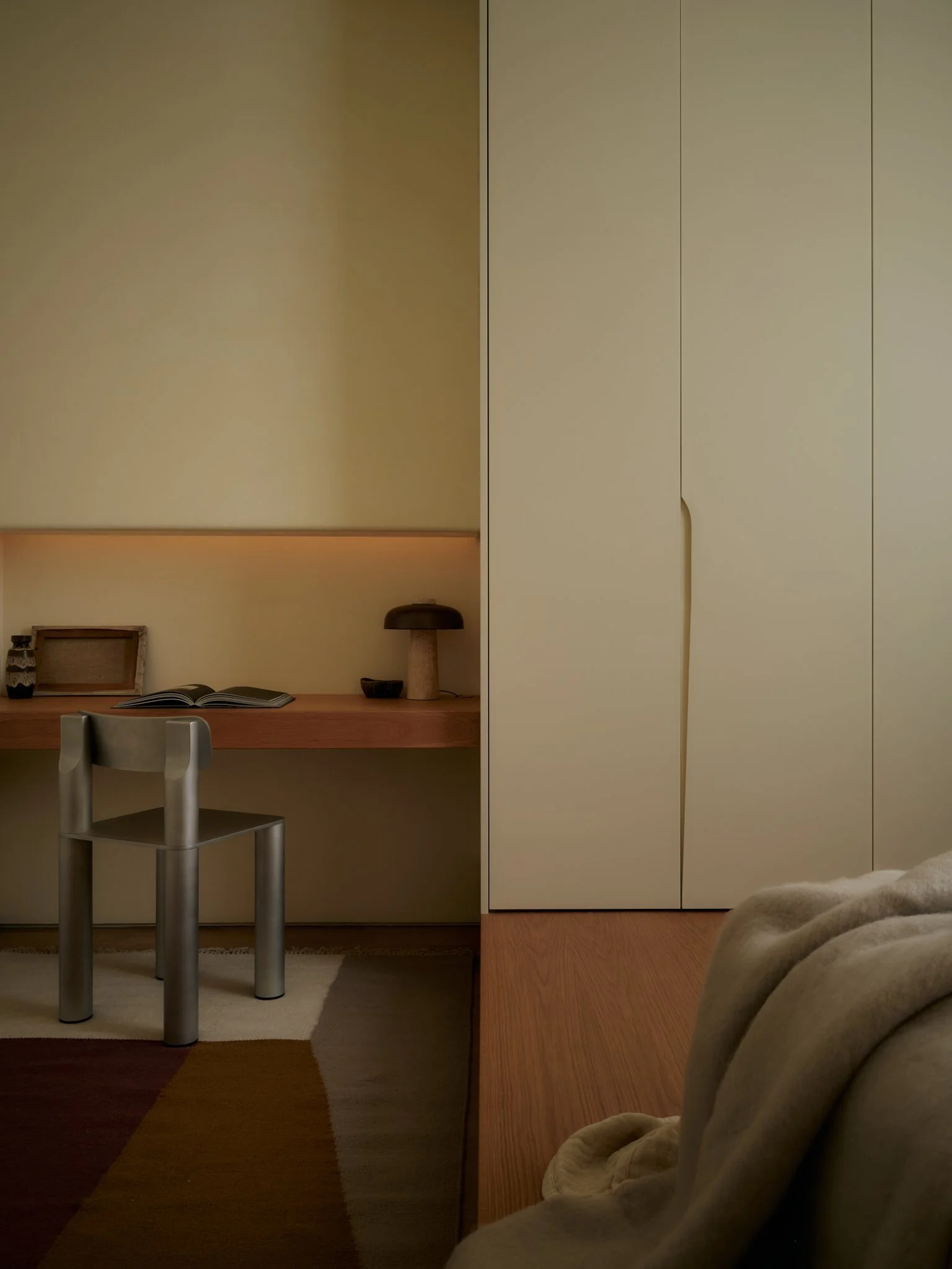 A bedroom with beige walls, a wooden desk with an open book, a framed picture, and decorative items, a black and beige stool, a beige closet door, and a bed partially visible in the foreground.