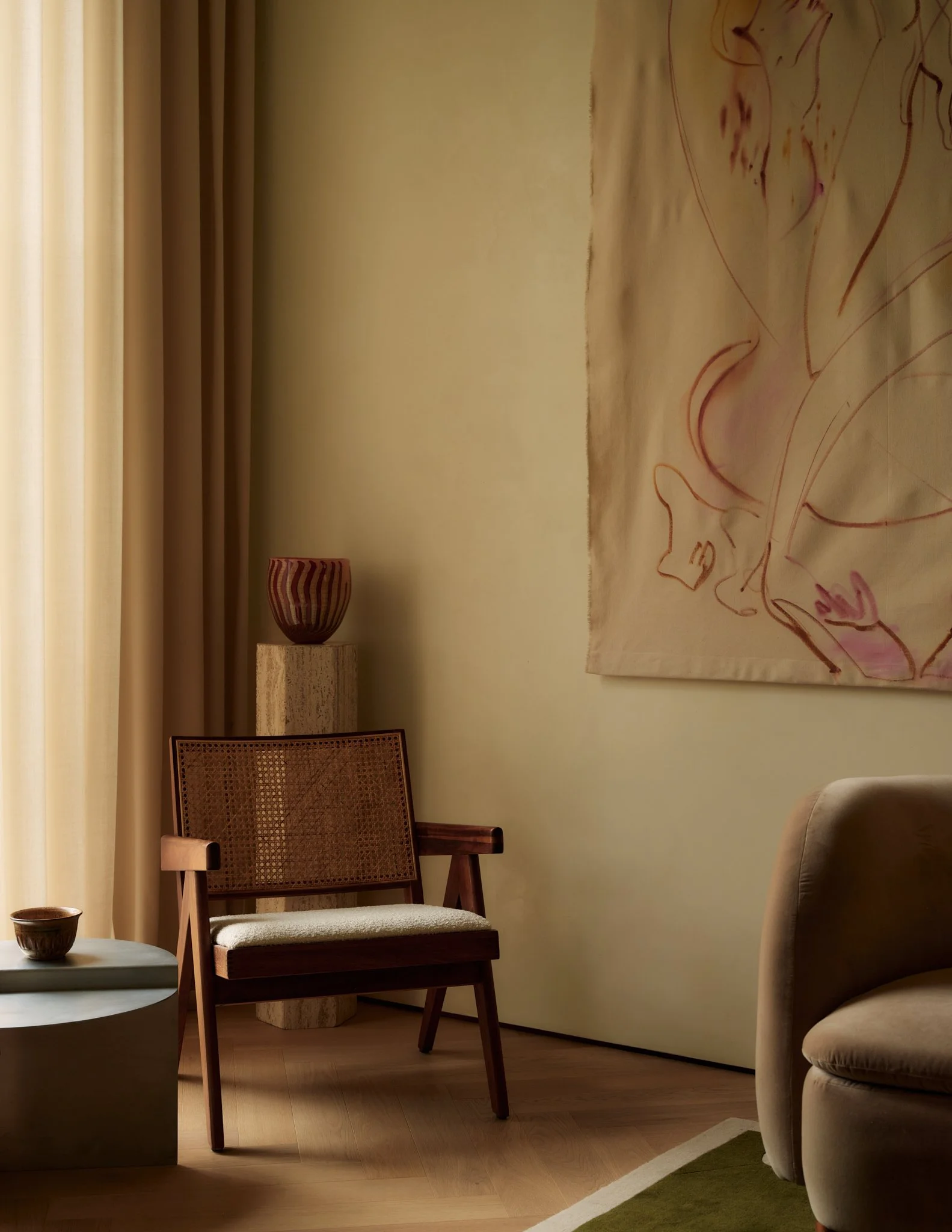 A cozy living room with a beige armchair, a wooden chair with woven backrest and a cushioned seat, a small table with a cup, and a large abstract profile artwork on the wall, in front of beige curtains and a pale yellow wall.