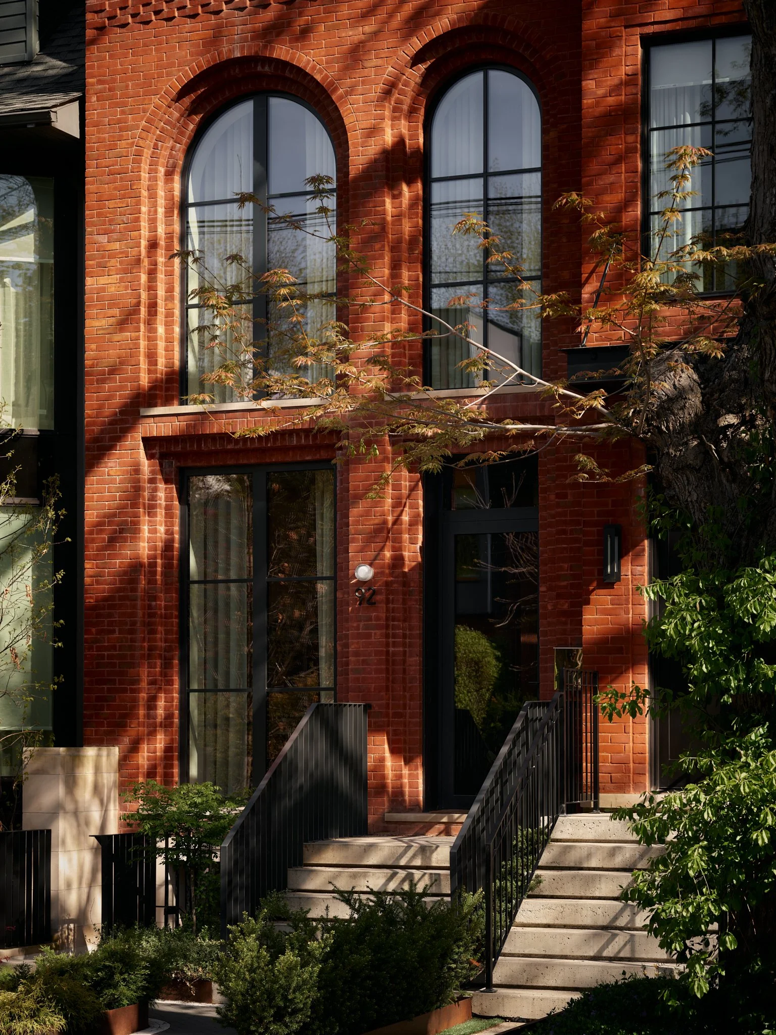 A brick building with large arched and rectangular windows and black metal stairs leading to the entrance, surrounded by green plants and trees.
