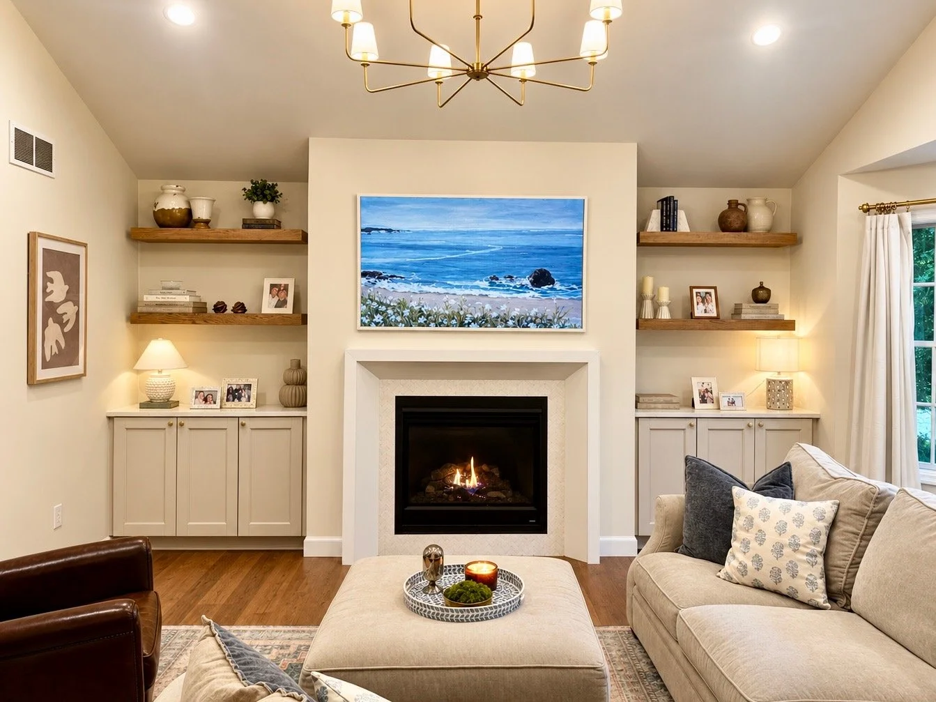 Living room with fireplace, beige sofa, throw pillows, coffee table with decorative items, wall art, built-in shelves with books and decor, chandelier, window with curtains, and a painting of a beach scene above the fireplace.