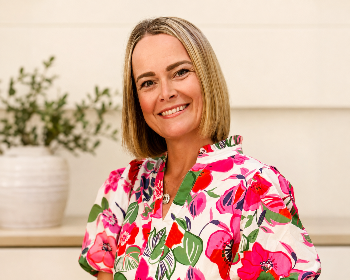A woman with shoulder-length blonde hair smiling at the camera, wearing a colorful floral-patterned top with pink, red, purple, and green shades, standing in front of a light-colored background with a potted plant.
