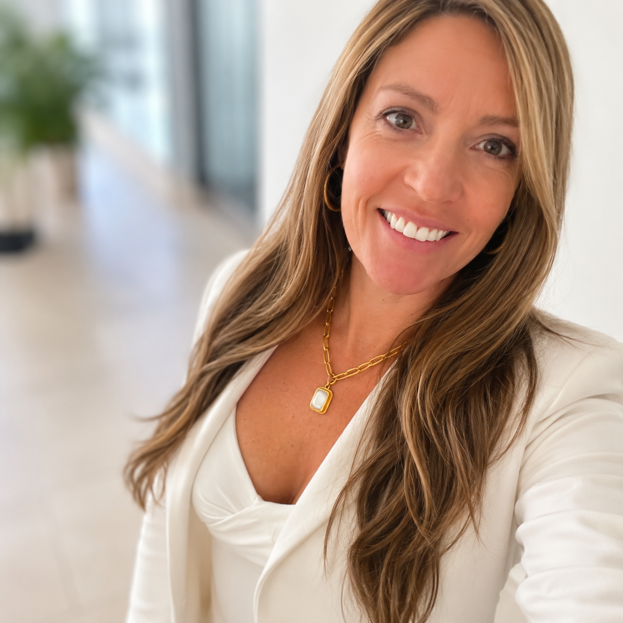 A woman with long light brown hair smiling, wearing a white blazer and a gold necklace, standing in a bright indoor setting.