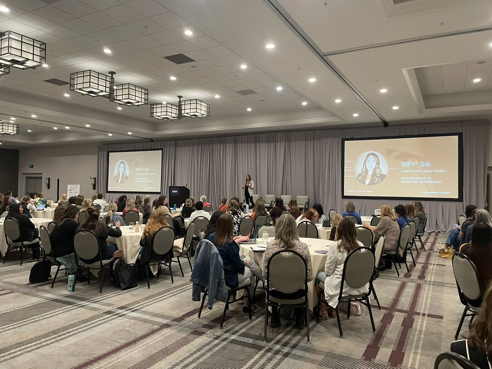 A speaker stands on stage in a large conference room, presenting to an audience seated at round tables. Two large screens display her presentation slides with a woman's illustration and text.