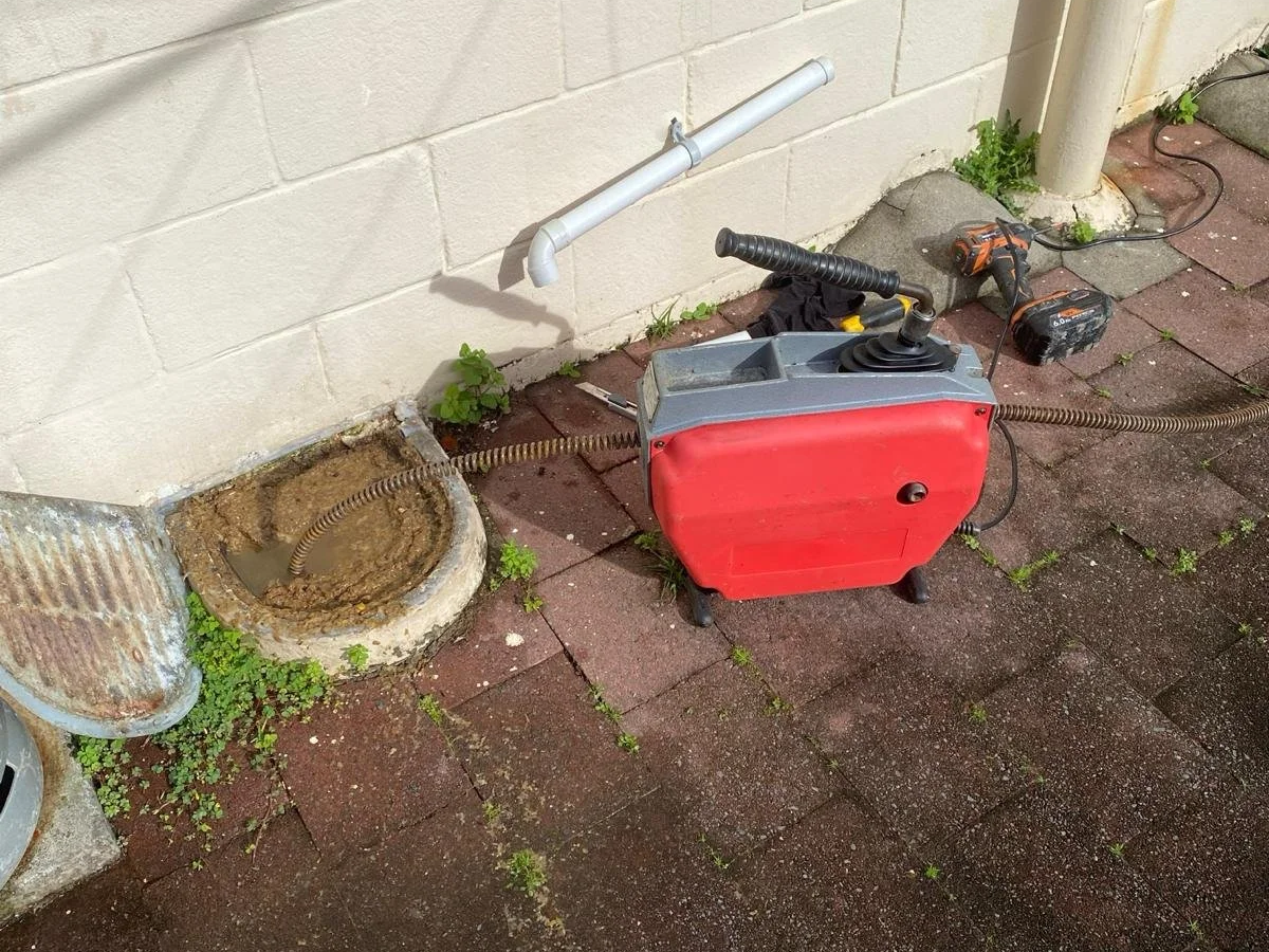 A sump pump is installed in a hole in the ground near a beige brick wall. The pump is red with a black handle and has a black control box. There is a white pipe attached to the wall and a black power drill nearby. Small green plants are growing around the area.