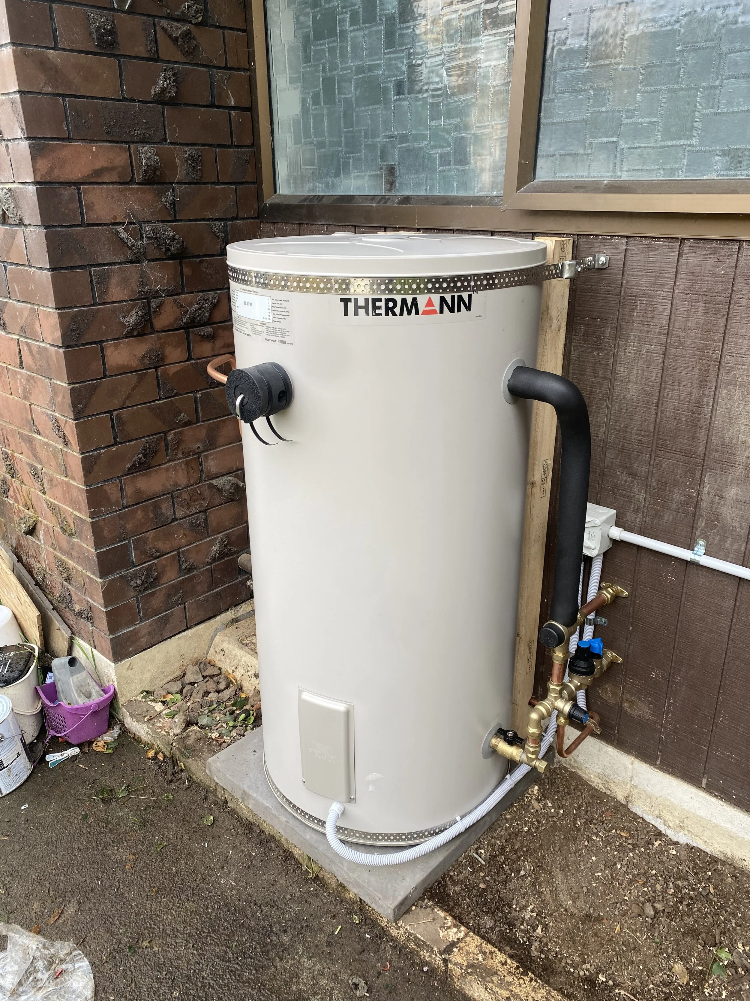 An outdoor hot water heater with pipes, a brick wall, a window, and gardening supplies on the ground.