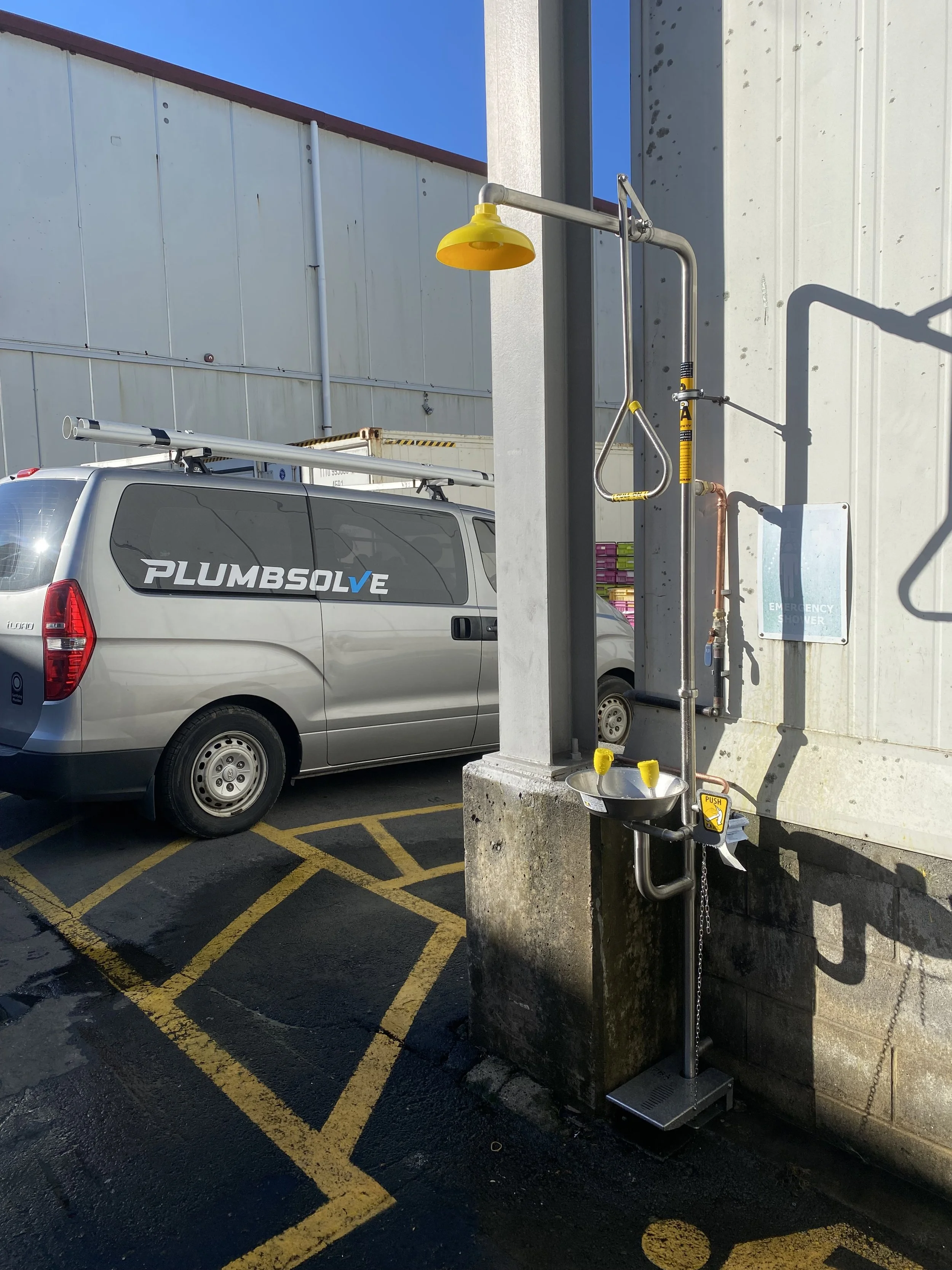 An outdoor scene with a white van labeled 'PLUMB SOLVE' parked near a concrete wall. Beside the wall, there is a hand washing station with a yellow soap dispenser, a foot pedal for water, and a metal frame, situated in a parking lot with painted yellow lines and a blue sky.