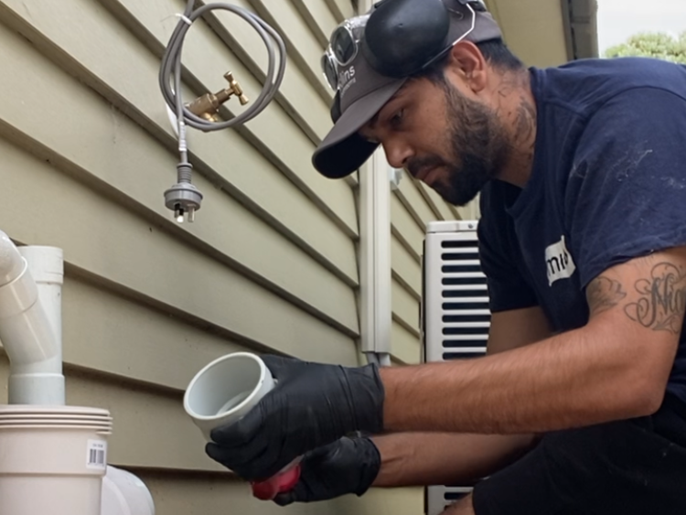 A man working outdoors on plumbing, wearing safety goggles, a cap, gloves, and a navy T-shirt, handling a pipe fitting.