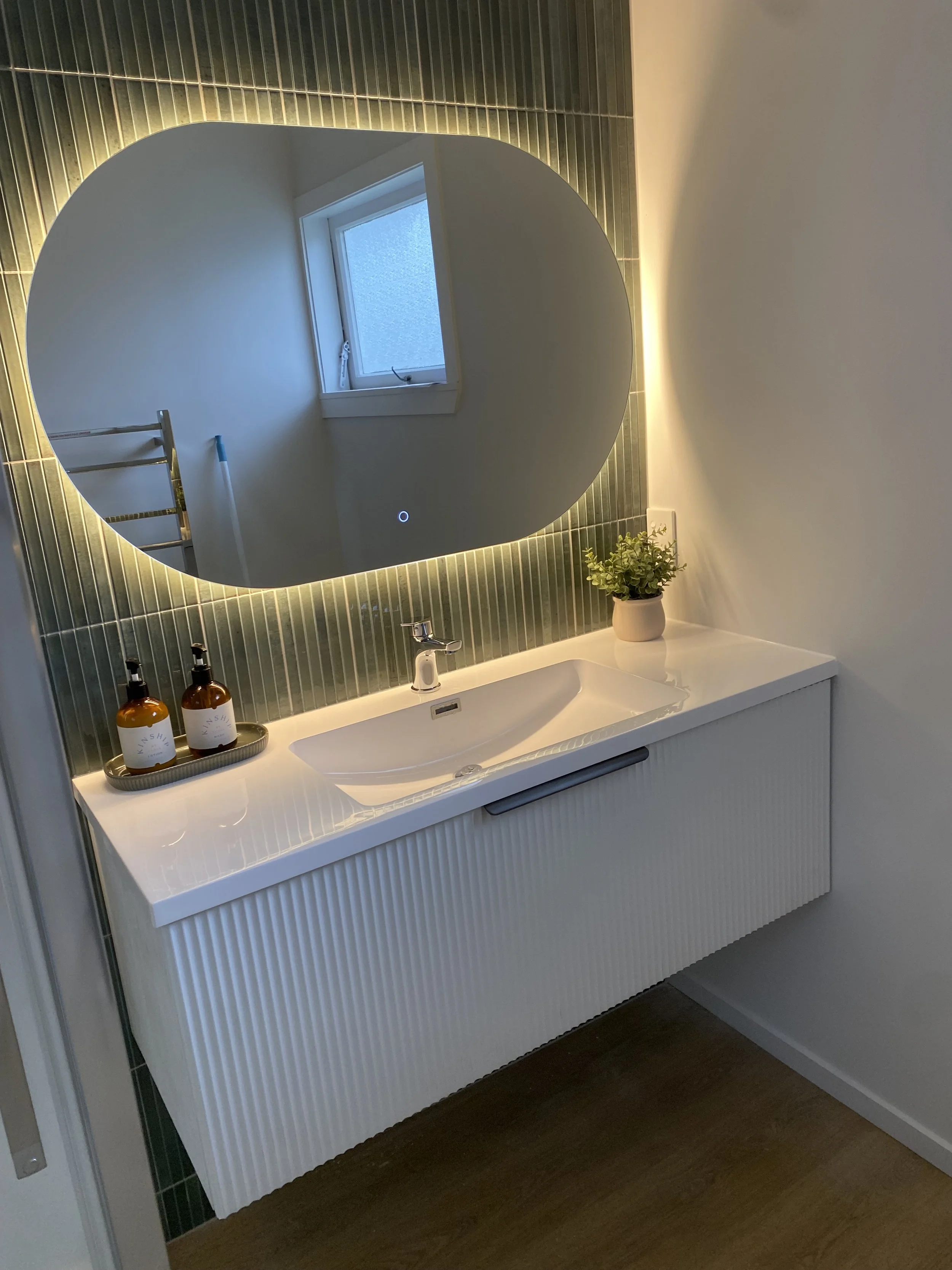 Bathroom vanity with a round mirror, two amber bottles on a tray, a small plant, a white sink, and dark wood flooring.