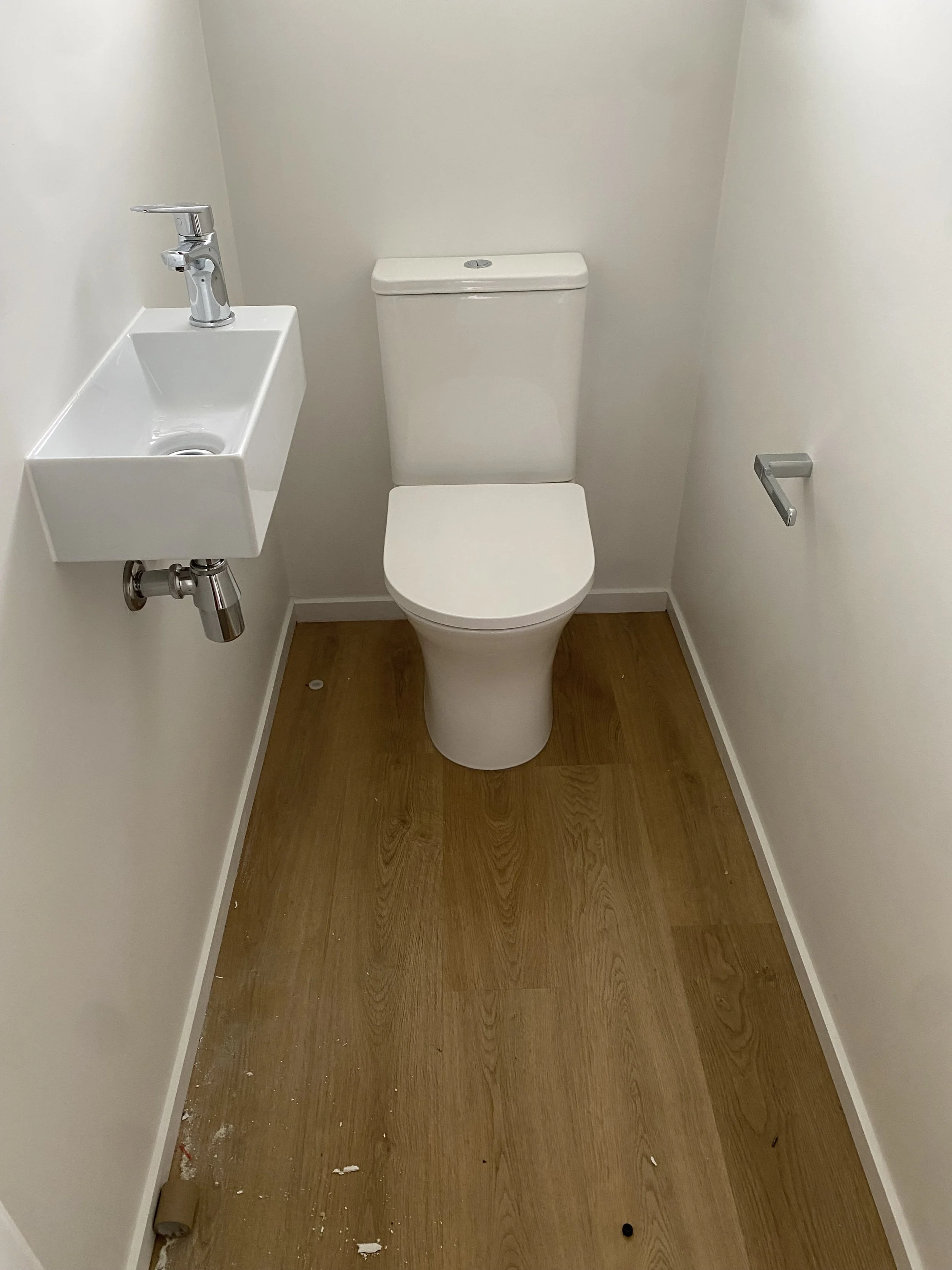 Small bathroom with a white toilet, a wall-mounted white sink with a chrome faucet, a toilet paper holder on the right wall, a roll of paper on the floor, and scattered debris on the wooden floor.