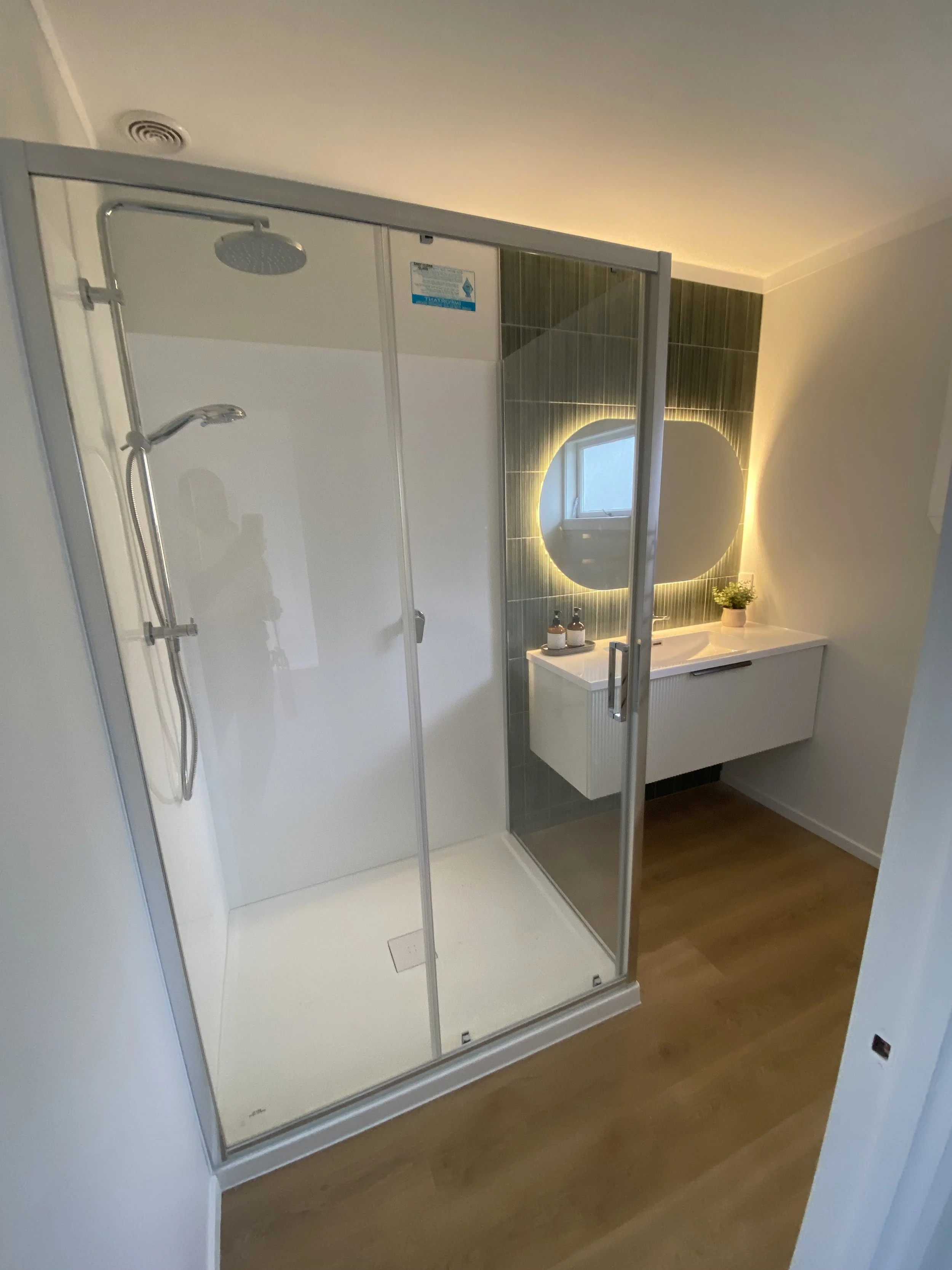 Modern bathroom with a glass shower enclosure, rain shower head, and a vanity with a round mirror and potted plant.