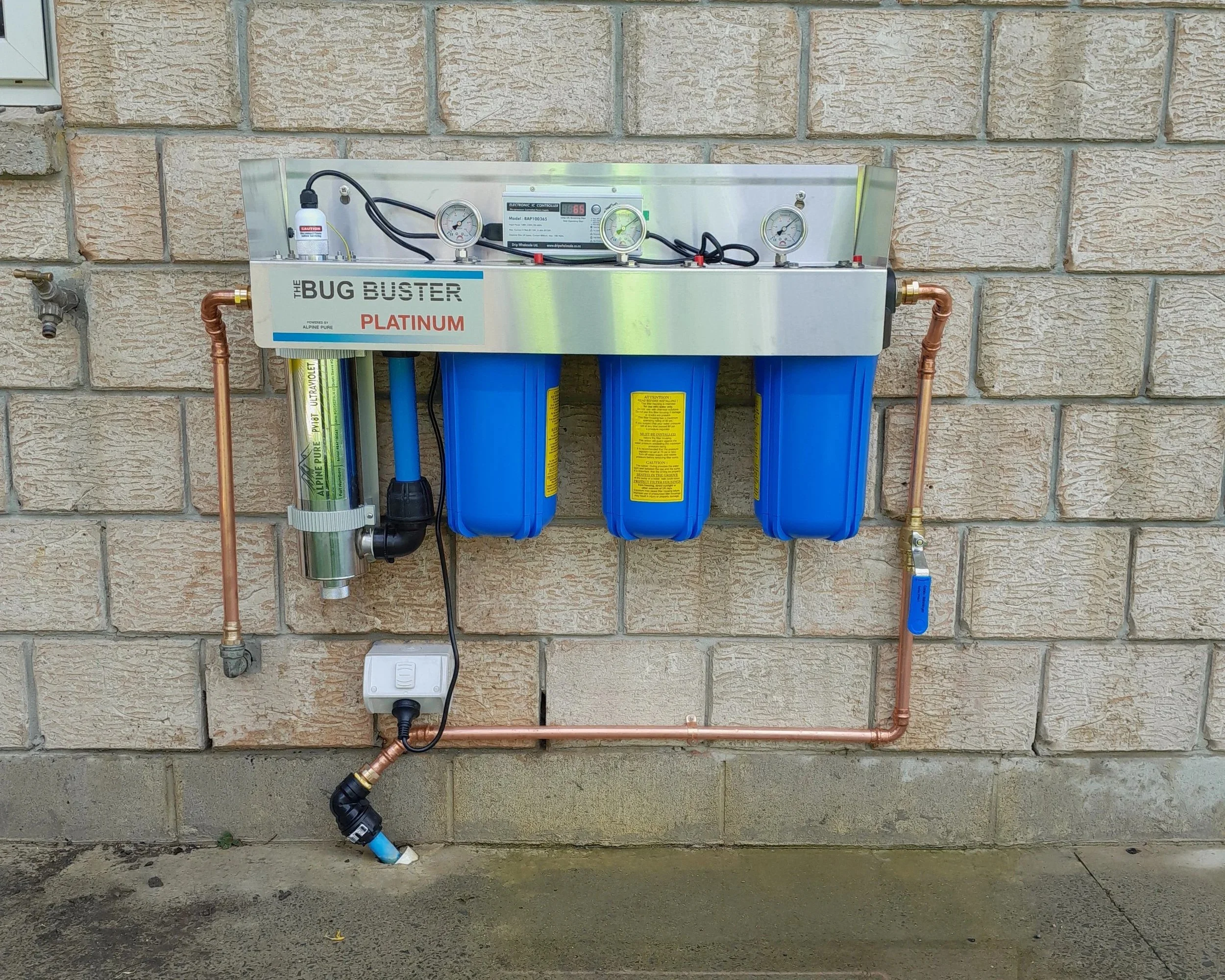 A water filter system mounted on a brick wall outside. It has three stage filter set up, copper pipes, gauges, and control units, with a faucet to the left.